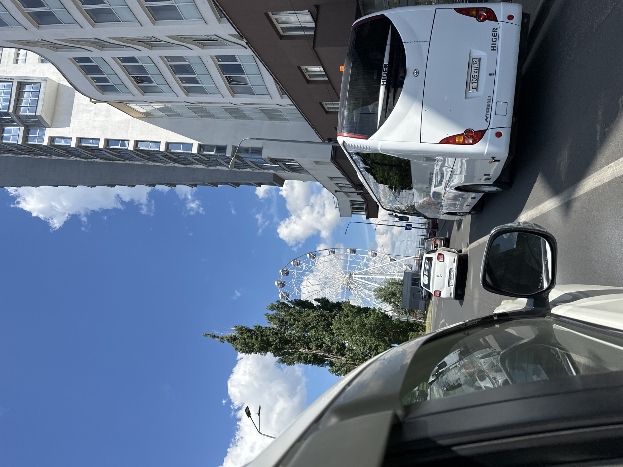 The image captures a street scene in Nizhny Novgorod, Russia, under a clear blue sky with scattered clouds. In the foreground, a white car's side is visible, indicating a perspective from within a vehicle. Ahead, a white bus with "HIGER" branding on its rear is traveling on the road. To the right, modern residential buildings with multiple floors and balconies dominate the skyline. To the left, a tall evergreen tree stands beside a Ferris wheel. Traffic signals and street lamps are also present. The overall scene depicts an urban environment with elements of leisure and everyday life.