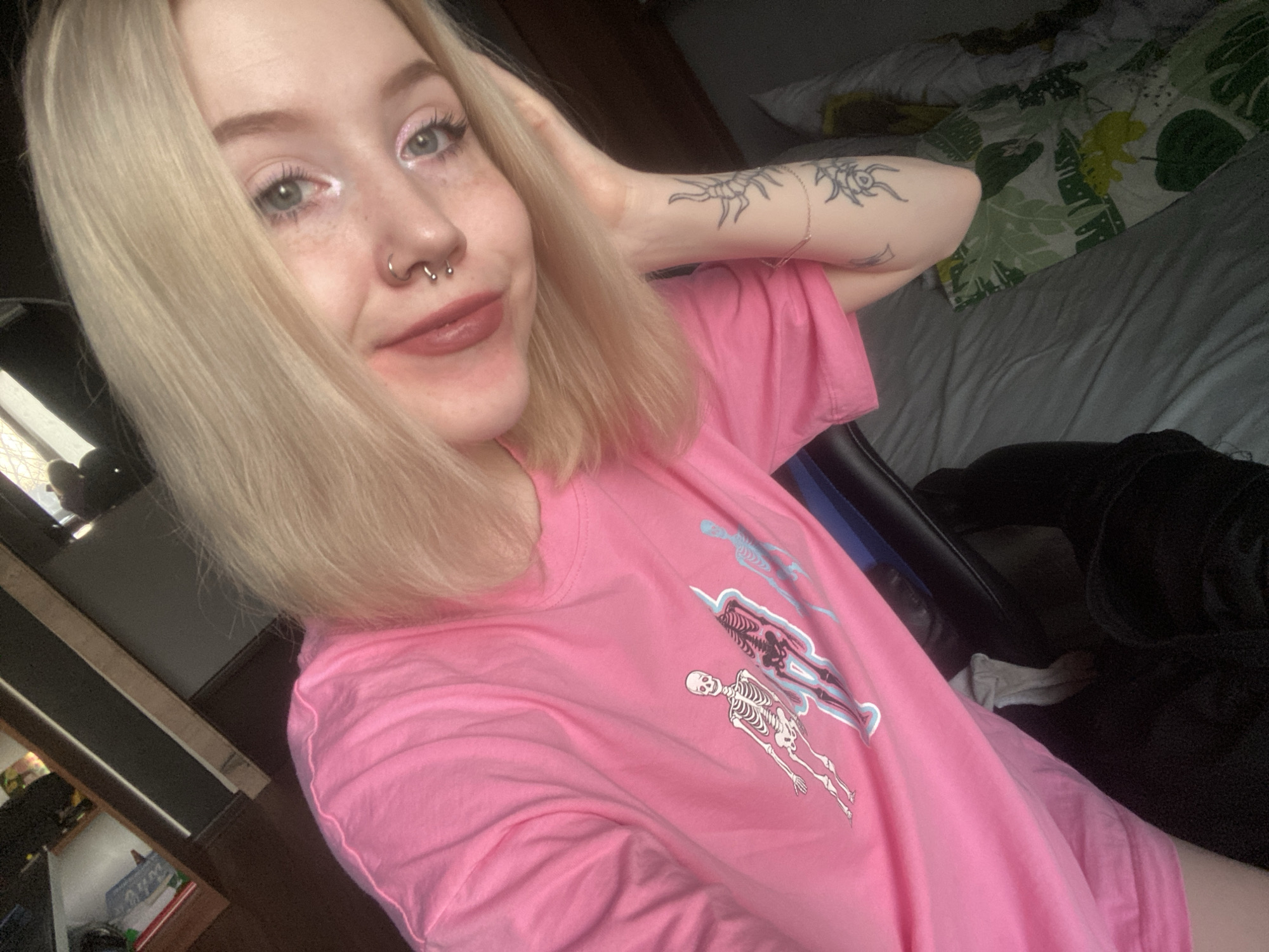 An individual with light blonde, shoulder-length hair, light eyes, and freckles is depicted in a medium shot, appearing to be a selfie. The individual has two septum piercings forming a circular barbell and is wearing reddish-brown lipstick and light pink and white eye makeup. Their left hand is positioned behind their head, displacing some hair. The left forearm displays multiple black ink tattoos, including a stylized arthropod design. The individual is wearing a bright pink t-shirt featuring a graphic design of multiple skeletons in white and light blue. The background shows an indoor setting, likely a bedroom, with a bed covered in grey sheets and a pillow with a green and yellow tropical leaf pattern visible to the right. A dark wooden furniture piece and a dark chair are partially visible to the left and lower right, respectively. The lighting suggests ambient indoor illumination. The location is Izhevsk, Russia.