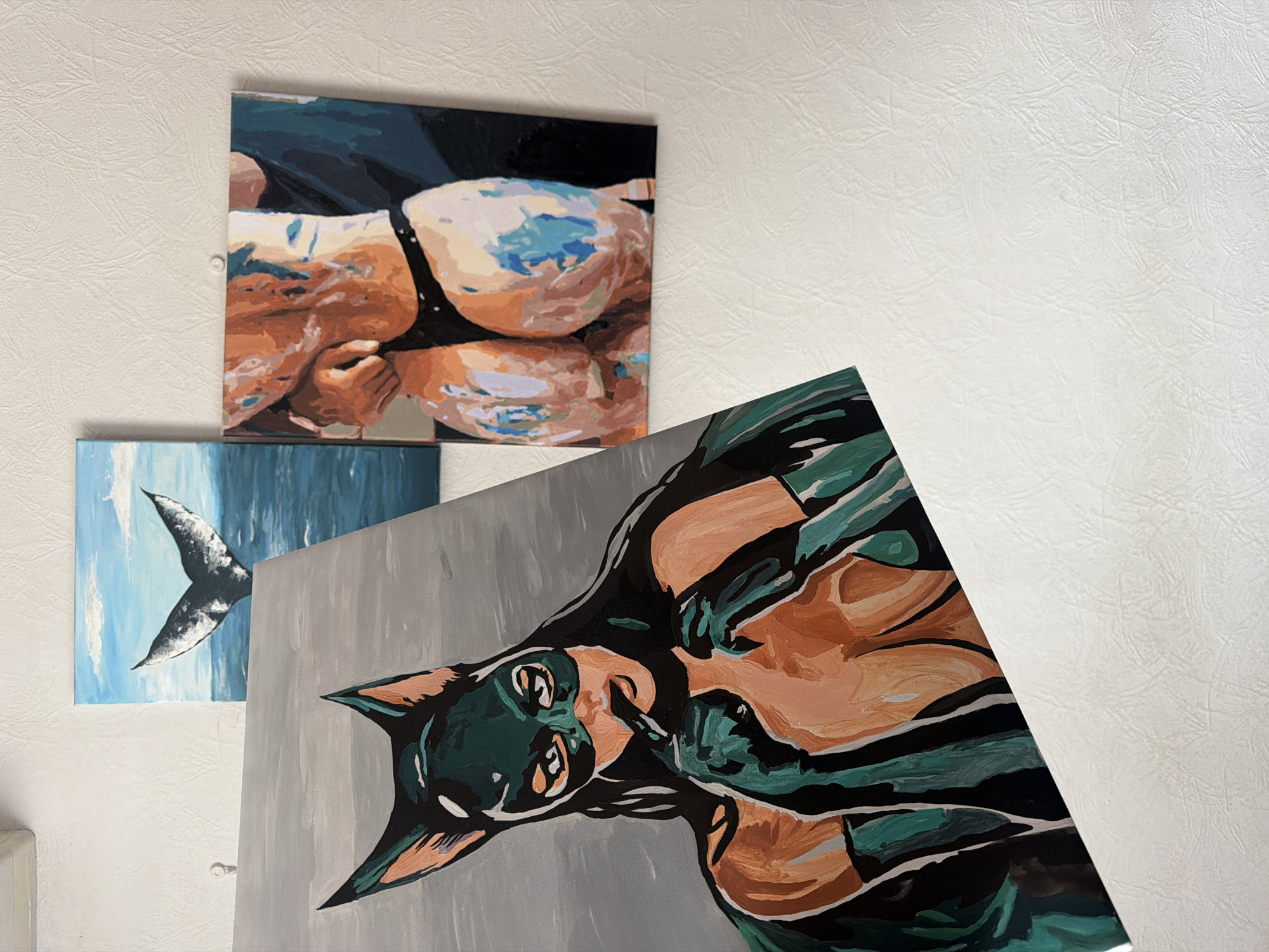 The image shows three paintings hanging on a white textured wall. The uppermost painting depicts a woman in a black swimsuit. Below it to the left is a painting of a whale tail emerging from the ocean. The painting in the foreground on the lower right appears to be a portrait of a Doberman Pinscher dog. The environment does not appear to show any features indicating the location is Moscow, Russia.