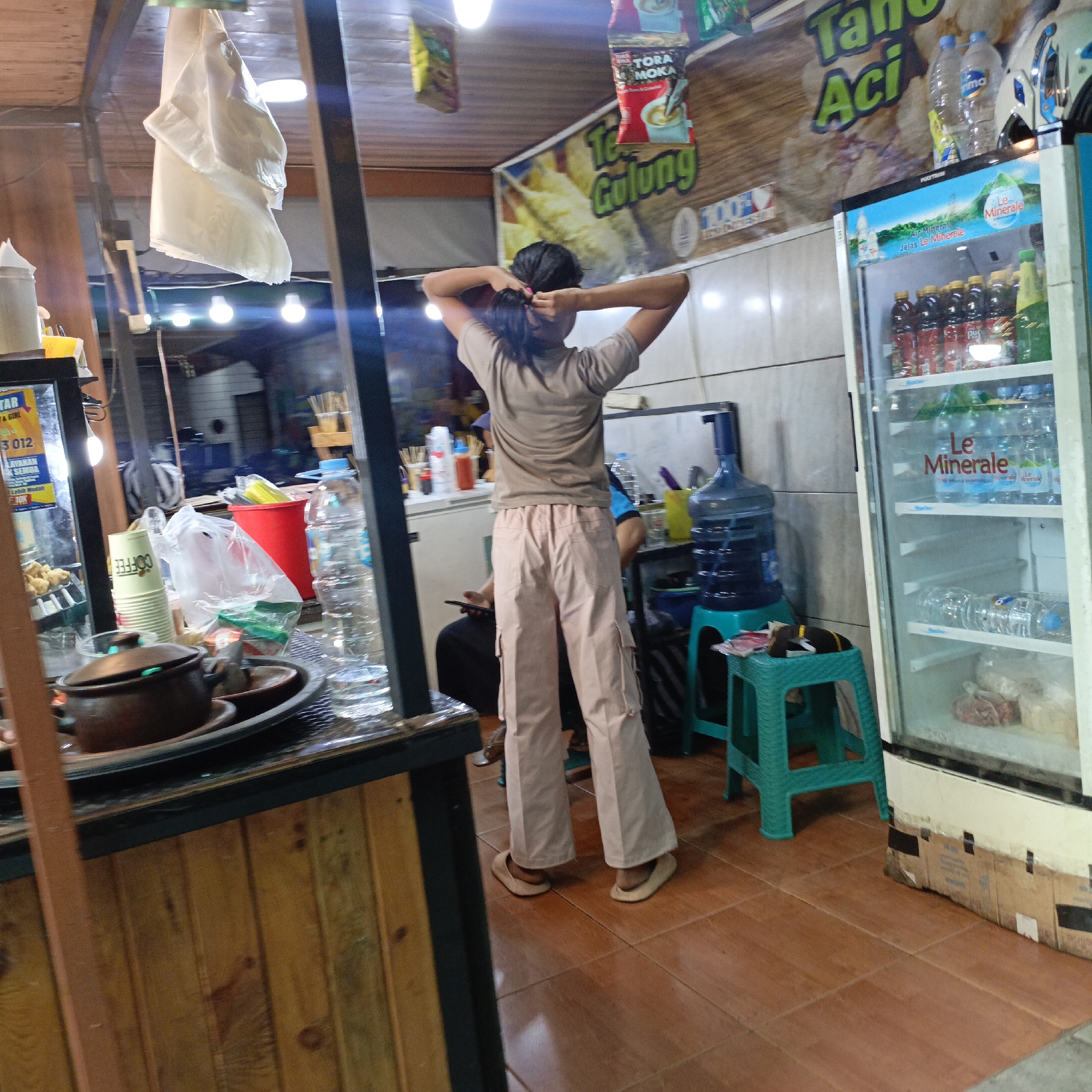 The image captures a street-level view of a small food stall or shop, likely in Salatiga, Indonesia, given the presence of Indonesian text on some of the signage. In the foreground, a wooden counter displays a dark-colored pot, some dishes, and various containers, including stacked paper cups with the word "COFFEE" printed on them, plastic bags, and bottles of water. To the right, a glass-fronted refrigerator displays bottled drinks, with brands like "Le Minerale" clearly visible. Adjacent to the refrigerator, a bright green plastic stool is placed near a large blue water dispenser.
A young person, viewed from behind, is standing in the middle ground, tying their dark hair up. They are wearing a light brown t-shirt and loose, light-colored cargo pants. To the left, a white fabric hangs from the ceiling, possibly a cloth cover or decoration. In the background, a banner with Indonesian text, including "Tahu Gulung," is displayed on a wooden wall. Other elements in the background suggest a busy, casual commercial environment with hanging packages of food products and more lighting fixtures. The overall impression is that of a small, informal business operating on a street or within a market area.