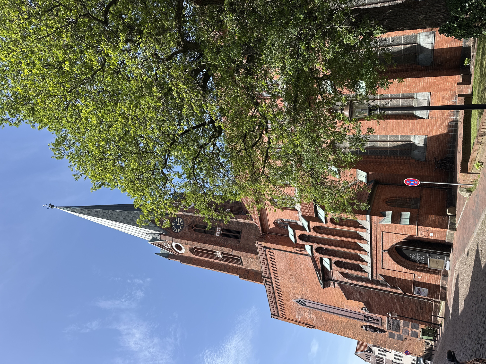 The image depicts St. Petri Church in Buxtehude, Germany. The church, constructed of red brick, features a tall steeple and an exterior clock. A large tree with green leaves partially obscures the view of the church. A street sign is visible in the foreground. The sky is blue with few clouds. The image appears to be taken from a low angle.