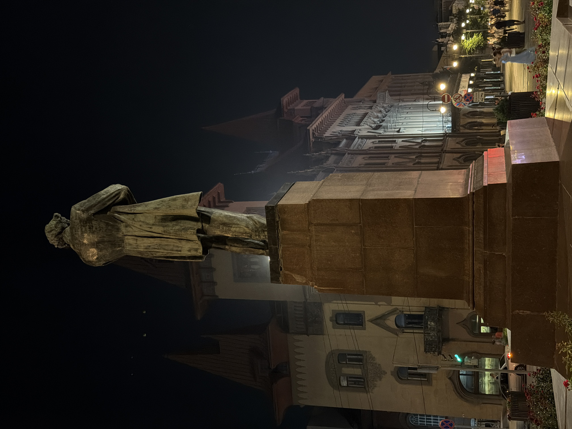 A night-time exterior shot showcases a bronze statue of a man from the back, standing on a large, multi-tiered stone pedestal. The statue is positioned in the foreground, dominating the lower half of the frame. Behind the statue, illuminated buildings with ornate architecture and pointed roofs are visible. To the left, a pale yellow building features arched windows and a balcony. To the right, a grander, lighter-colored building with many windows and intricate detailing lines the street. A row of streetlights with ornate fixtures illuminate the sidewalk, where several people are walking. A traffic light with a green signal is visible to the left of the statue. In the lower foreground, red flowers bloom. The scene is characteristic of a public square in the city of Saratov, Russia, likely in the vicinity of the monument to Nikolai Chernyshevsky, given the statue's style and the architectural elements reminiscent of buildings in the city center.