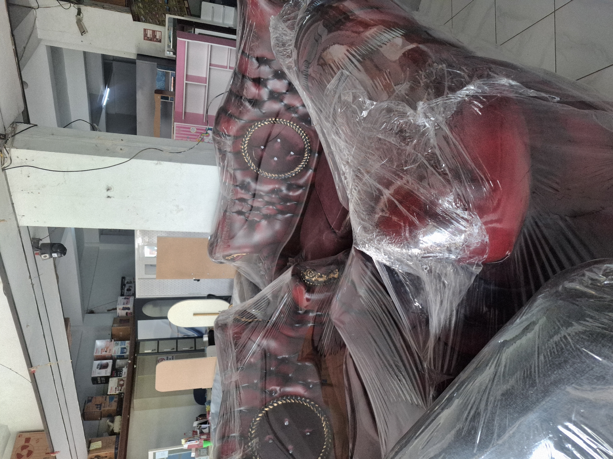 The image shows a store interior in Banjarmadu, Indonesia, with several pieces of furniture, primarily sofas and chairs, wrapped in clear plastic. The furniture is upholstered in a dark red, possibly leather, material and features decorative tufting and gold trim. In the background, there are shelves stocked with various boxed items, likely home appliances or electronics, and other furniture items like a pink bookshelf. The environment appears to be a retail space, possibly a furniture store or a warehouse. The lighting is somewhat dim, with visible fluorescent lights overhead and natural light filtering in from unseen sources. The floor is tiled. There are no people visible in the image.