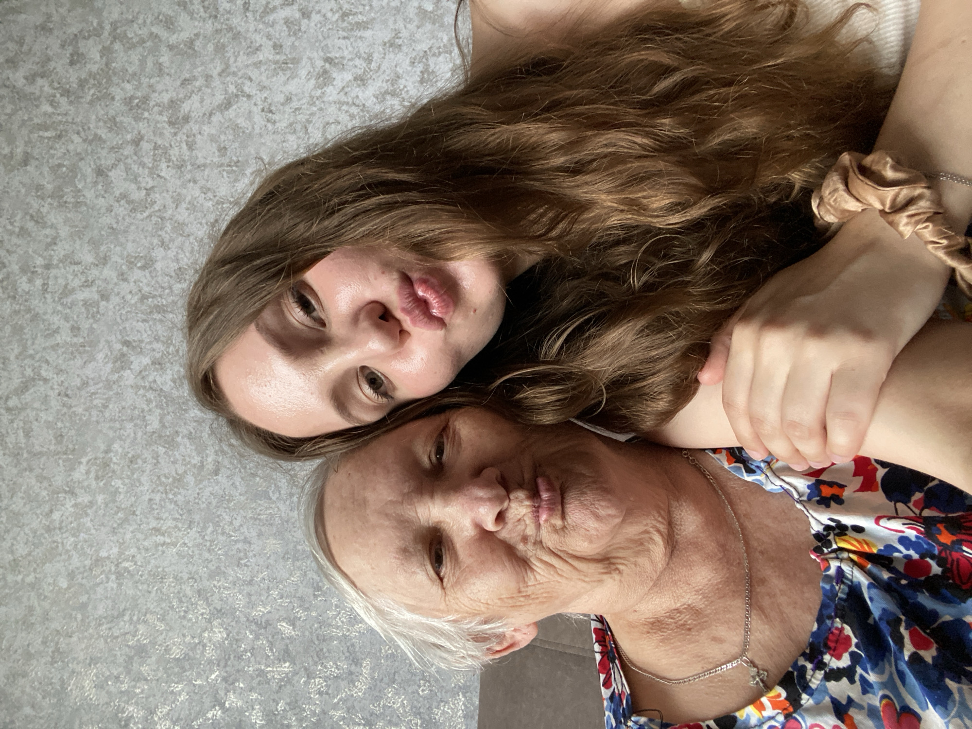 A close-up selfie taken indoors in Марковский, Russia, depicts two females, an elderly woman and a younger woman, positioned in the foreground against a textured grey wall. Both individuals are looking directly at the camera with their lips pursed in a "kissy face" expression.
The elderly woman occupies the left side of the frame, displaying fair skin with numerous facial wrinkles and short, swept-back grey hair. She is wearing a multi-colored top with a floral or abstract pattern and a thin silver chain necklace with a small pendant. A dark brown surface, likely a sofa, is visible behind her.
The younger woman is positioned to the right, partially behind the elderly woman, with long, wavy, light brown hair flowing over her shoulder. She has fair skin and is wearing a light-colored ribbed top. Her right hand is placed affectionately on the elderly woman's left arm, and a light brown satin scrunchie is visible on her left wrist.