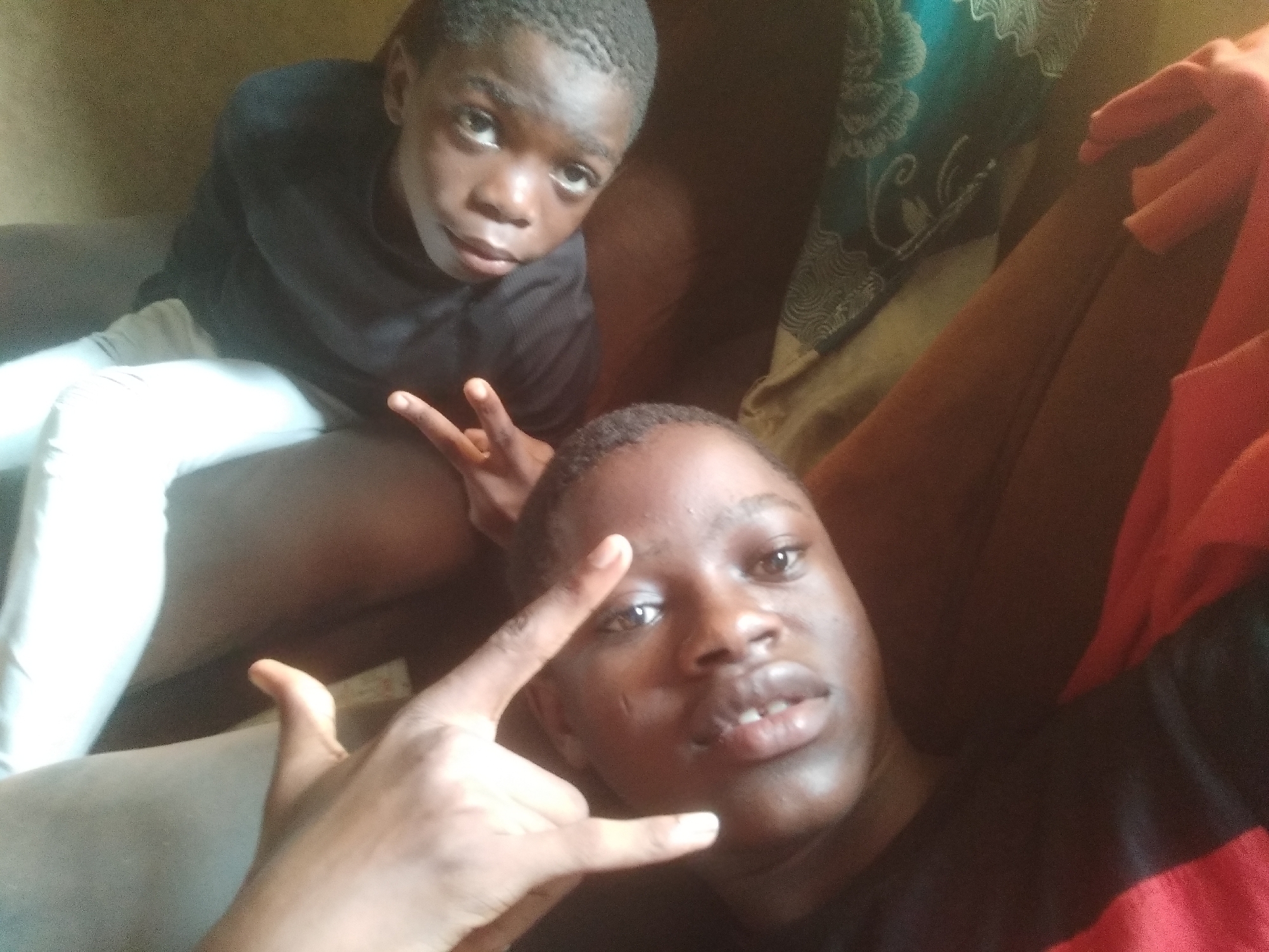 Two young individuals are captured in what appears to be a casual indoor selfie. The person in the foreground is positioned closer to the camera, with their head and upper torso visible. This individual has dark skin, short dark hair, and is wearing a black garment with a red stripe or panel on the right side. They are making a "rock on" or "devil horns" hand gesture with their left hand, holding it up near their face. Their expression is neutral.
Behind and slightly to the left, another young person is seated, appearing a bit further back and less in focus. This individual also has dark skin and short dark hair, and is wearing a black long-sleeved top and white pants. They are making a "peace sign" hand gesture with their right hand, positioned near their leg. Their gaze is directed towards the camera.
The setting appears to be a living space, likely on or near a sofa, with brown upholstery visible. A patterned fabric, possibly a cushion or blanket with a teal and light design, is seen in the upper right background. The lighting is soft, suggesting an indoor environment, possibly with indirect natural light or artificial lighting. No text is visible in the image. The overall mood is relaxed and casual, characteristic of a shared moment between friends or family members.