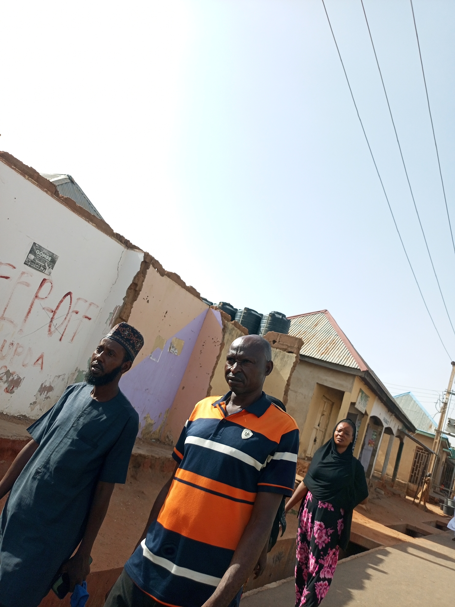 Three individuals are walking on a street in what appears to be a built-up area. The first individual, a male, is wearing a dark blue robe and a patterned cap. The second individual, also a male, is wearing a striped orange and blue polo shirt. The third individual, a female, is wearing a black hijab and a floral skirt. The environment includes buildings, some of which appear to be in a state of disrepair. Graffiti is visible on one of the buildings. Utility poles and wires are also present. Based on the prompt's instructions, the location is presumed to be in the city of Tempure, Nigeria.