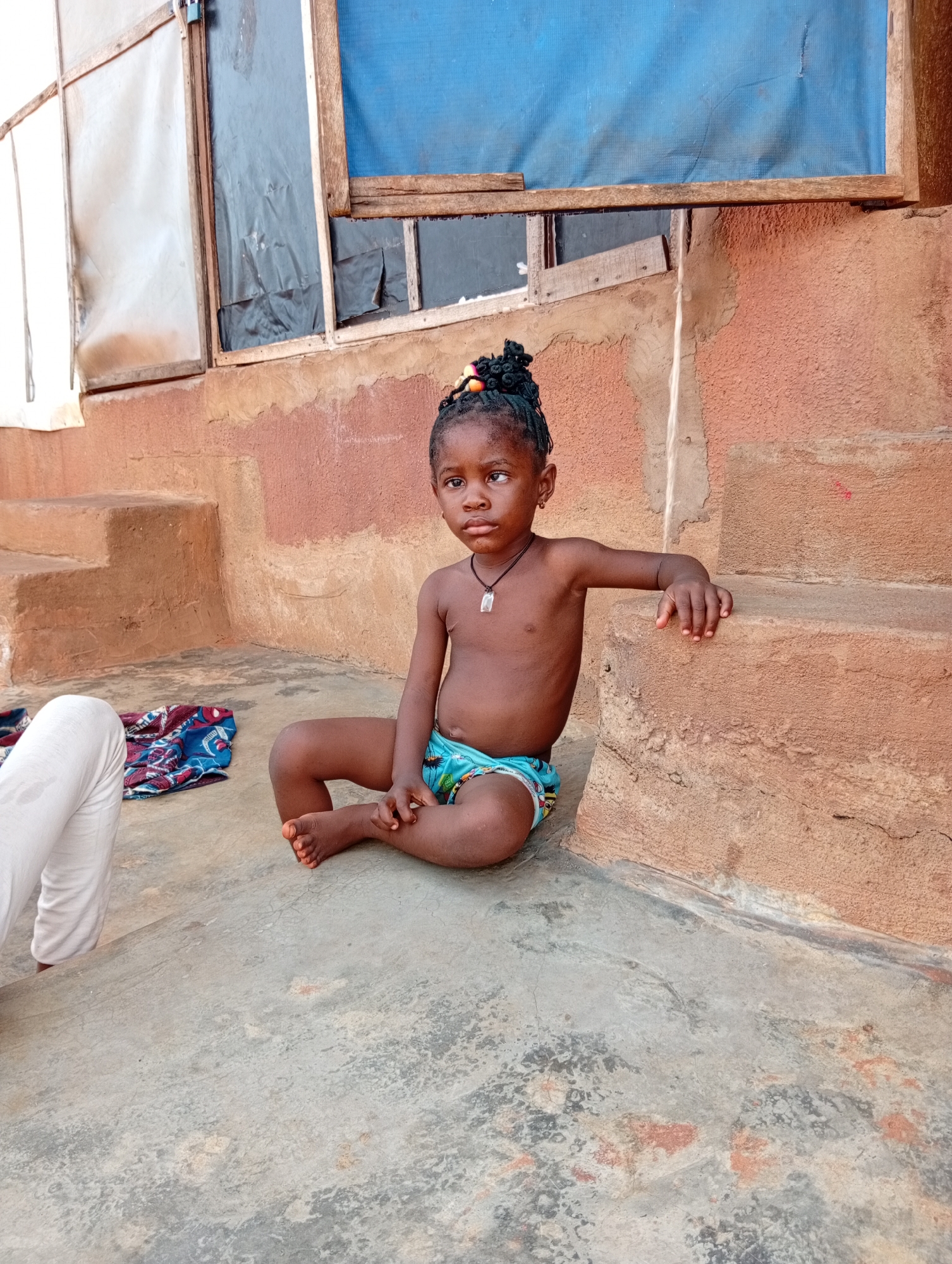 A young Black child is seated outdoors on a concrete surface, likely in Ogidi, Nigeria. The child, possibly a girl, has dark skin, braided hair styled with colorful bands, and wears light blue patterned briefs. She has a dark cord necklace with a clear pendant and small earrings. Her left arm rests on a low, broad, reddish-brown concrete step, and she looks intently to the left of the frame with a serious expression.
To the child's immediate left, part of an adult's leg in white trousers is visible, along with a folded piece of colorful, possibly ankara, patterned fabric on the ground. The background features a rustic building with textured, reddish-brown walls and several concrete steps. Above the steps, there are rough wooden frames enclosing openings that appear to be makeshift windows. These openings are covered with various materials: a translucent white sheet, black plastic sheeting, and a blue mesh screen on the far right.
The scene is lit by clear daylight, suggesting a bright day. The simple construction materials of the building indicate a humble or rural setting. The child appears to be resting or observing her environment. No specific activity beyond sitting is evident, and there is no visible text in the image.