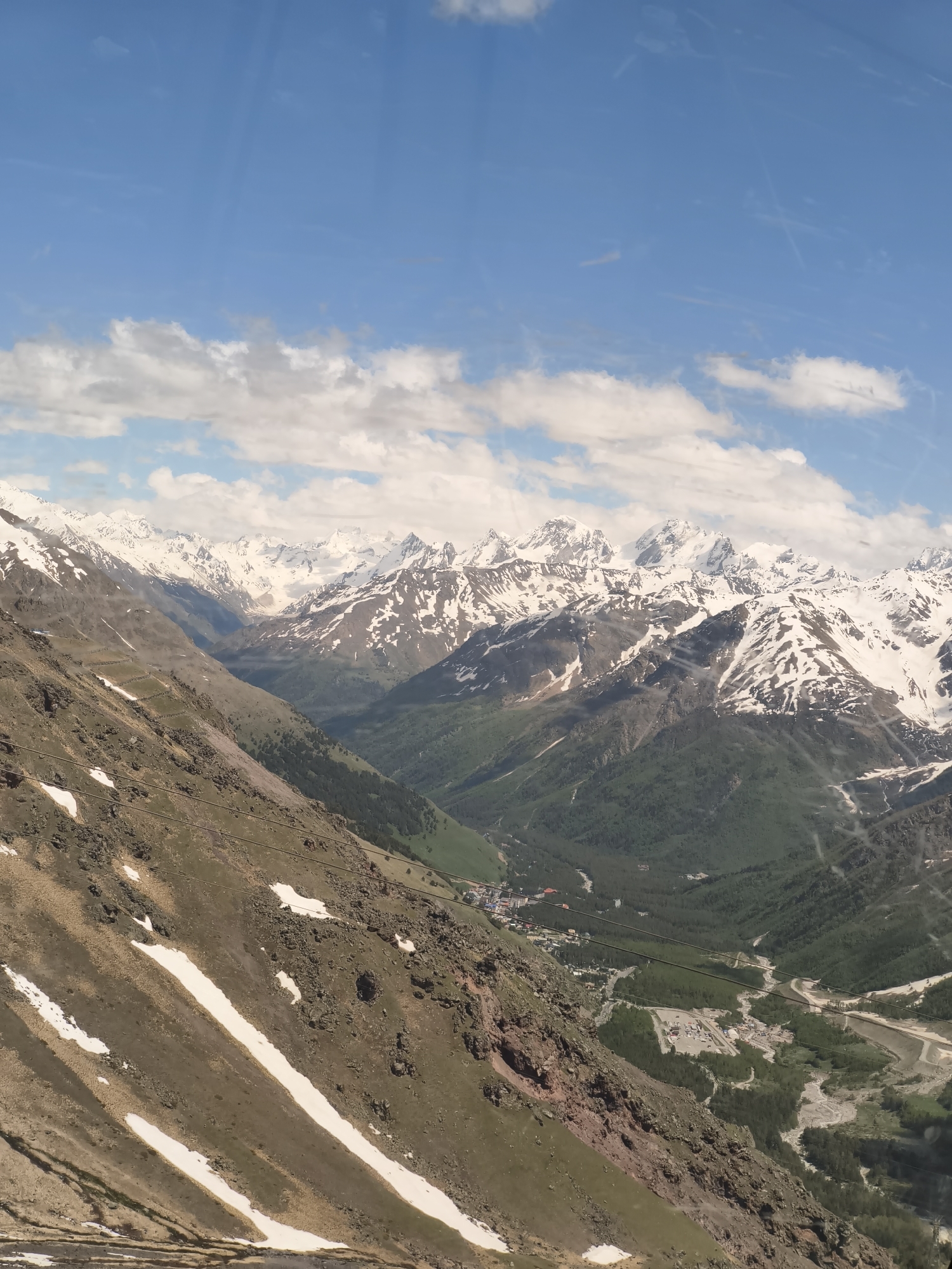 The image captures a breathtaking panoramic view of a mountain valley, likely in or near Terskol, Russia. The foreground is dominated by a steep, rocky mountainside with patches of snow scattered across its brown and green terrain. Several parallel wires, possibly from a ski lift or cable car, traverse this slope diagonally.
Further into the scene, the valley opens up, revealing a lush green landscape dotted with trees. A winding river or streambed carves through the valley floor, its path marked by lighter colored sand and gravel. In the distance, nestled within the valley, a small town or settlement can be observed with buildings and infrastructure.
The background is filled with majestic, snow-capped mountain peaks that stretch across the horizon. Their rugged slopes and white summits create a dramatic contrast with the vibrant green of the valley below. The sky above is a clear blue, adorned with scattered white clouds, some of which appear wispy and elongated. The overall impression is one of a vast, natural landscape, highlighting the grandeur of the Caucasus Mountains.