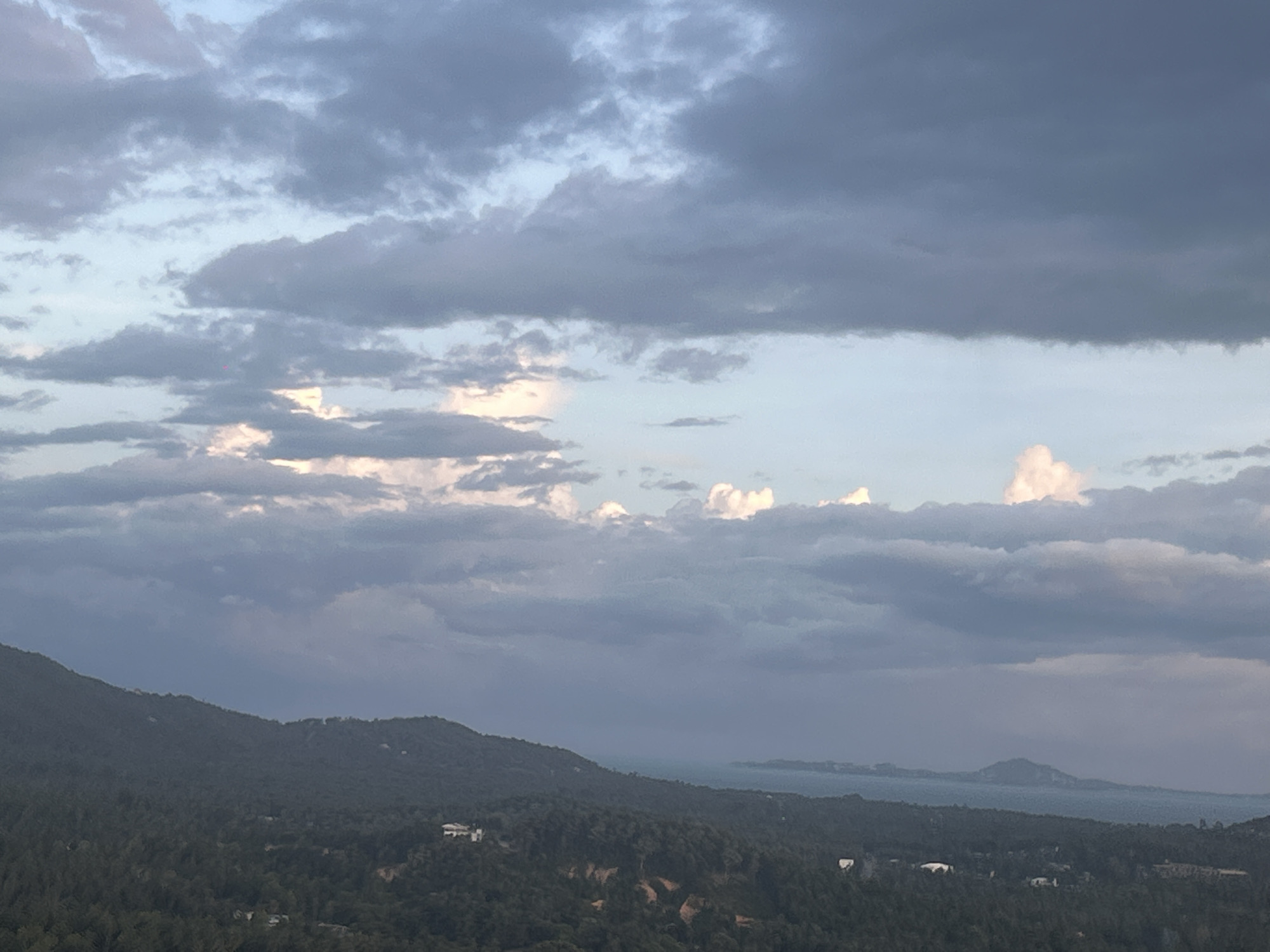 The image depicts a landscape view near Baan Hin Kong, Thailand, under a cloudy sky. A mountainous region covered with dense trees is visible in the foreground. The hills are dark green and suggest a lush forest. In the background, there is a body of water, possibly the sea, with a few islands or landmasses. The sky is dominated by dark gray clouds, with some areas of lighter, brighter clouds. The overall lighting is dim, indicating either early morning, late evening, or overcast conditions. The scene gives a sense of remoteness and natural beauty.