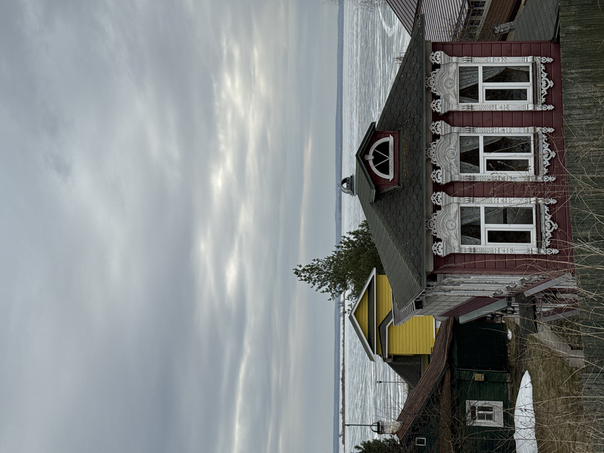 The image shows a row of buildings in Ново-Никольское, Russia, with a body of water in the background. The buildings are various colors, including maroon, yellow, and green. The maroon building features decorative window frames. The sky is overcast. The body of water appears to be partially frozen or covered with ice. Bare trees and branches are visible in the foreground.