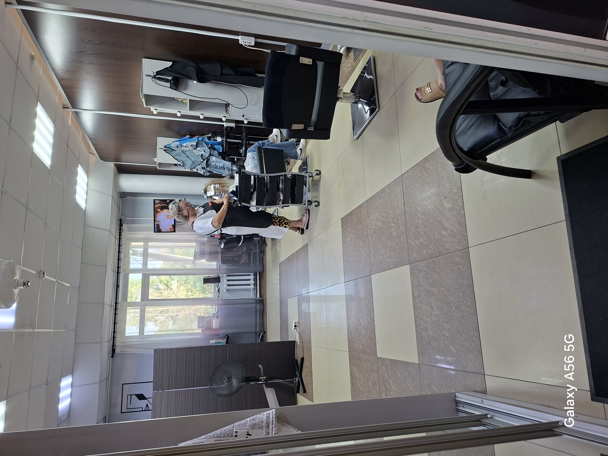 This image depicts the interior of a hair salon. In the foreground, a black salon chair is positioned on a tiled floor. To the left, a large mirror or wardrobe unit is partially visible. In the midground, a woman wearing an apron and leopard-print pants is attending to a client, who is seated and has her hair being worked on. A rolling trolley with various hair products and tools is next to the client. Further in the background, a standing fan is visible near a window with white curtains. The walls are a mix of light colors and wood paneling. The scene appears to be set in a well-lit, functional salon environment. No specific indicators of the city of Chita, Russia are present in the image itself.