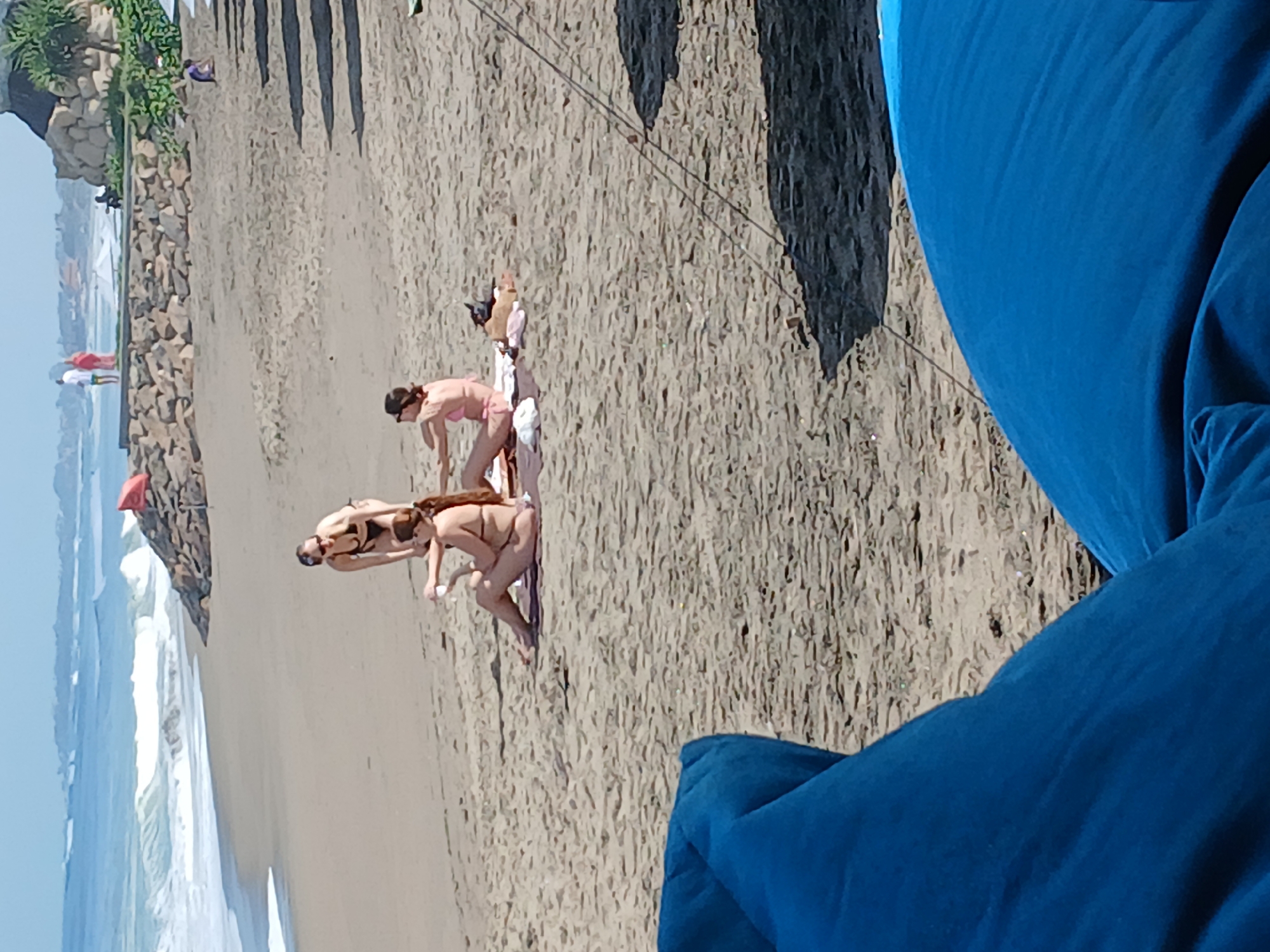 A group of three women are on a sandy beach, possibly in Bali, Indonesia, given the tropical vegetation and architecture. The women are wearing bikinis and appear to be socializing or relaxing. One woman is standing and talking to the other two who are seated on towels on the sand. The ocean is visible in the background with waves breaking on the shore, and a rocky embankment runs along the coastline. Further in the background, there are buildings and people walking. The foreground is occupied by the edge of what appears to be a blue lounger or beanbag chair. The sky is clear and the sun is shining, indicating a bright, sunny day.