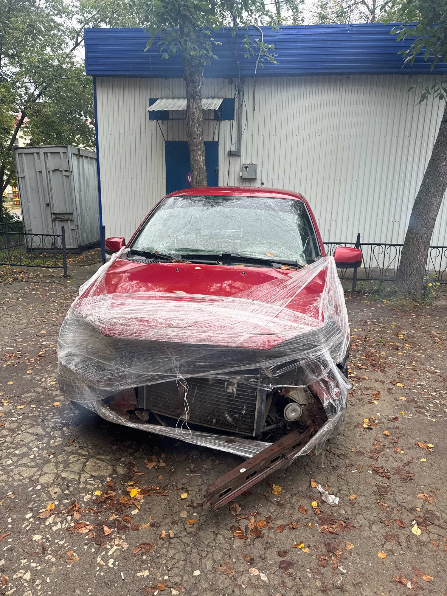 The image depicts a red sedan, its front end heavily damaged and wrapped in clear plastic sheeting. The vehicle is parked on an asphalt surface covered with fallen leaves. In the background, a light-colored building with a blue corrugated metal roof is visible, along with a metal fence and trees. The overall scene suggests an urban environment, possibly a parking lot or street in Ulyanovsk, Russia, given the context provided. No people are present in the image.