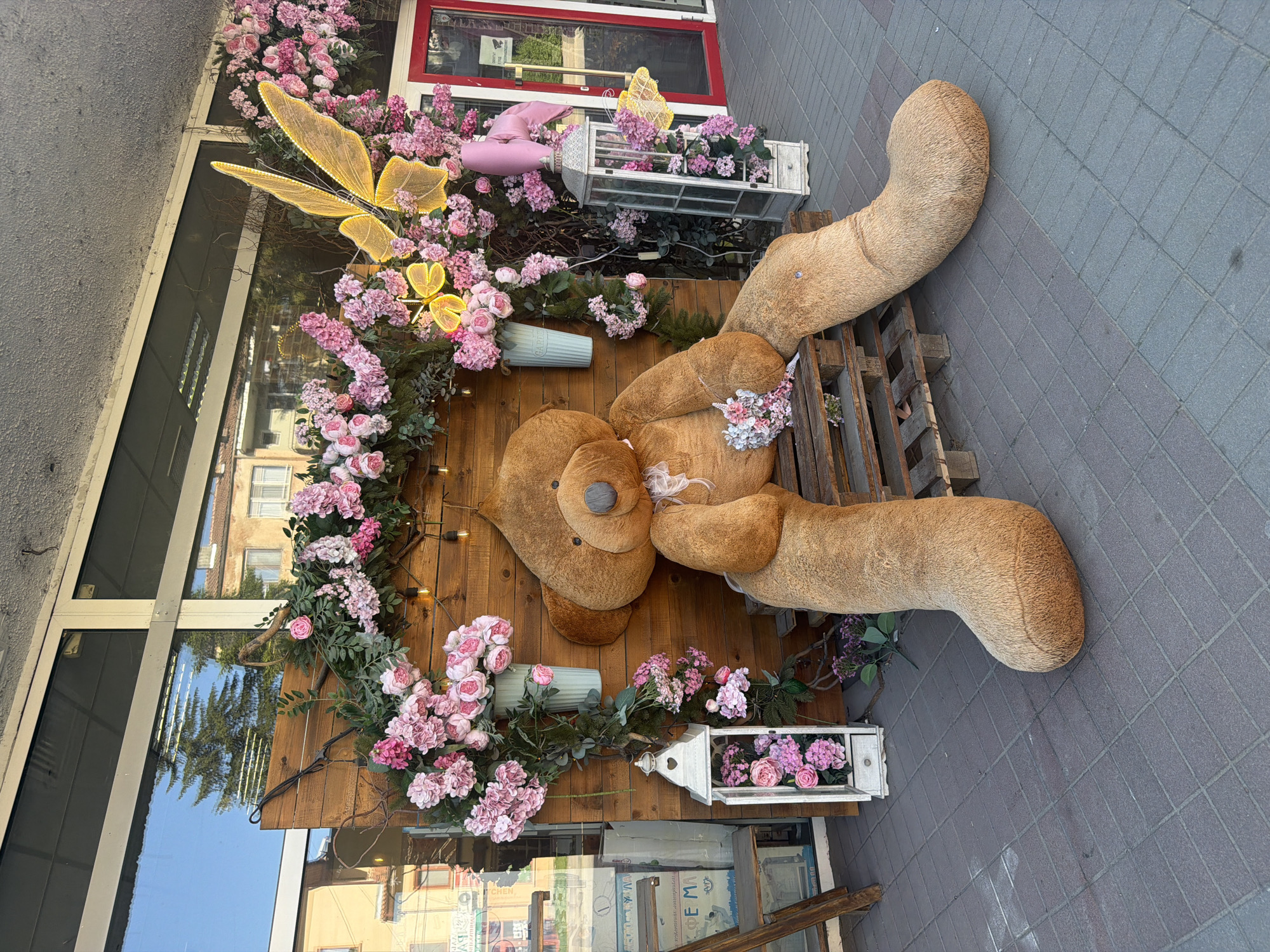 The image shows a large teddy bear decoration on a wooden board adorned with pink flowers and lit by fairy lights. The bear is resting on a wooden pallet placed on a gray tiled surface. The scene is outside a building with a glass facade, reflecting buildings and the sky. The location is likely in Lukovit, Bulgaria, as indicated by the prompt. Additional decorations include a glowing butterfly, small lanterns, and flower pots. The overall ambiance suggests a festive or decorative setting.