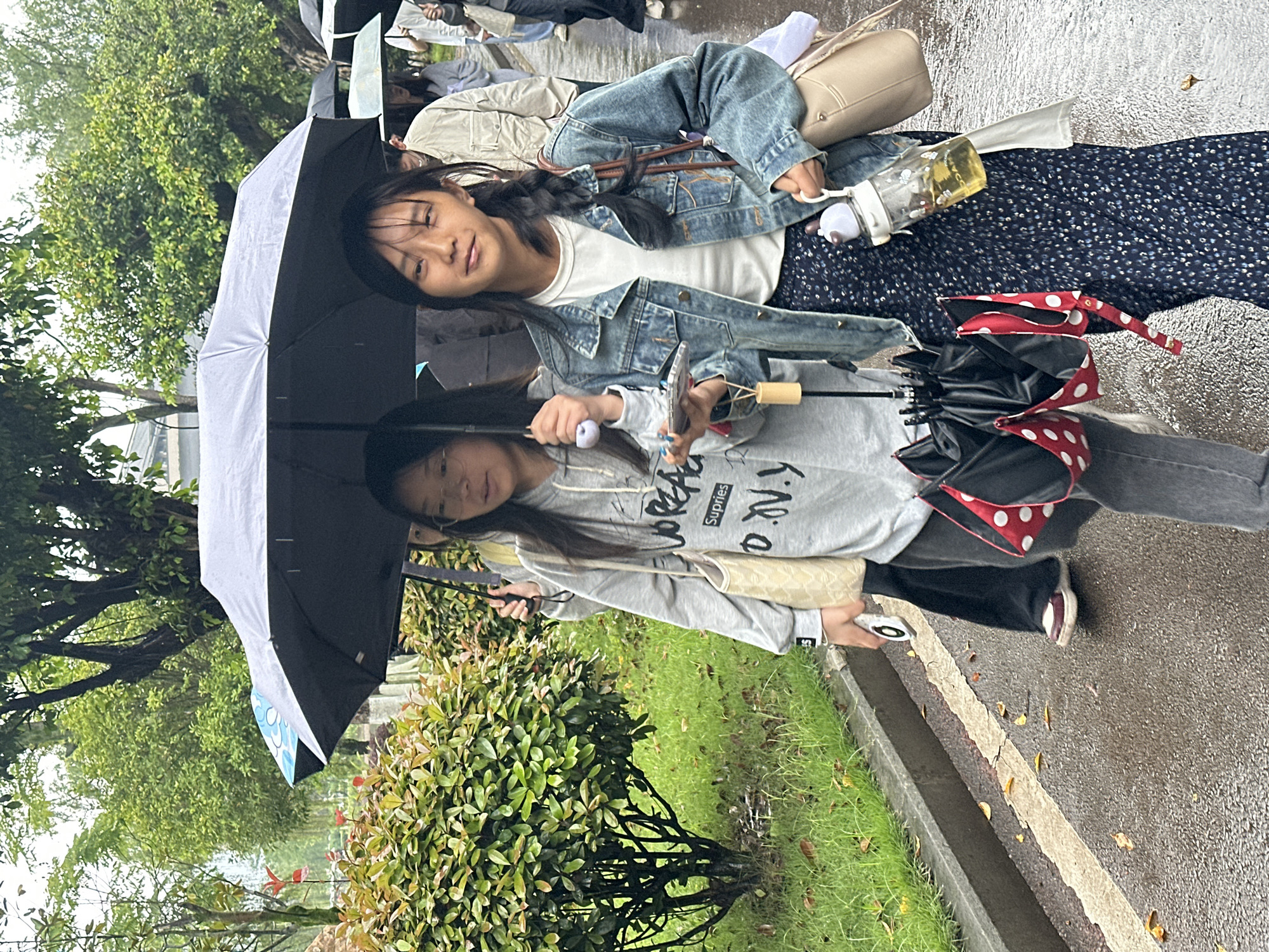 Two young women, both holding open umbrellas, walk along a paved street beside a curb in an outdoor environment characterized by green foliage and trees, likely Jiangjin, China. One woman wears a denim jacket over a white shirt and a dark patterned skirt, while the other is dressed in a grey hooded sweatshirt and dark pants. One of the umbrellas has red and white polka dot accents. Both women face the camera. The street appears wet, suggesting it has been raining. Additional people and umbrellas are visible in the background.