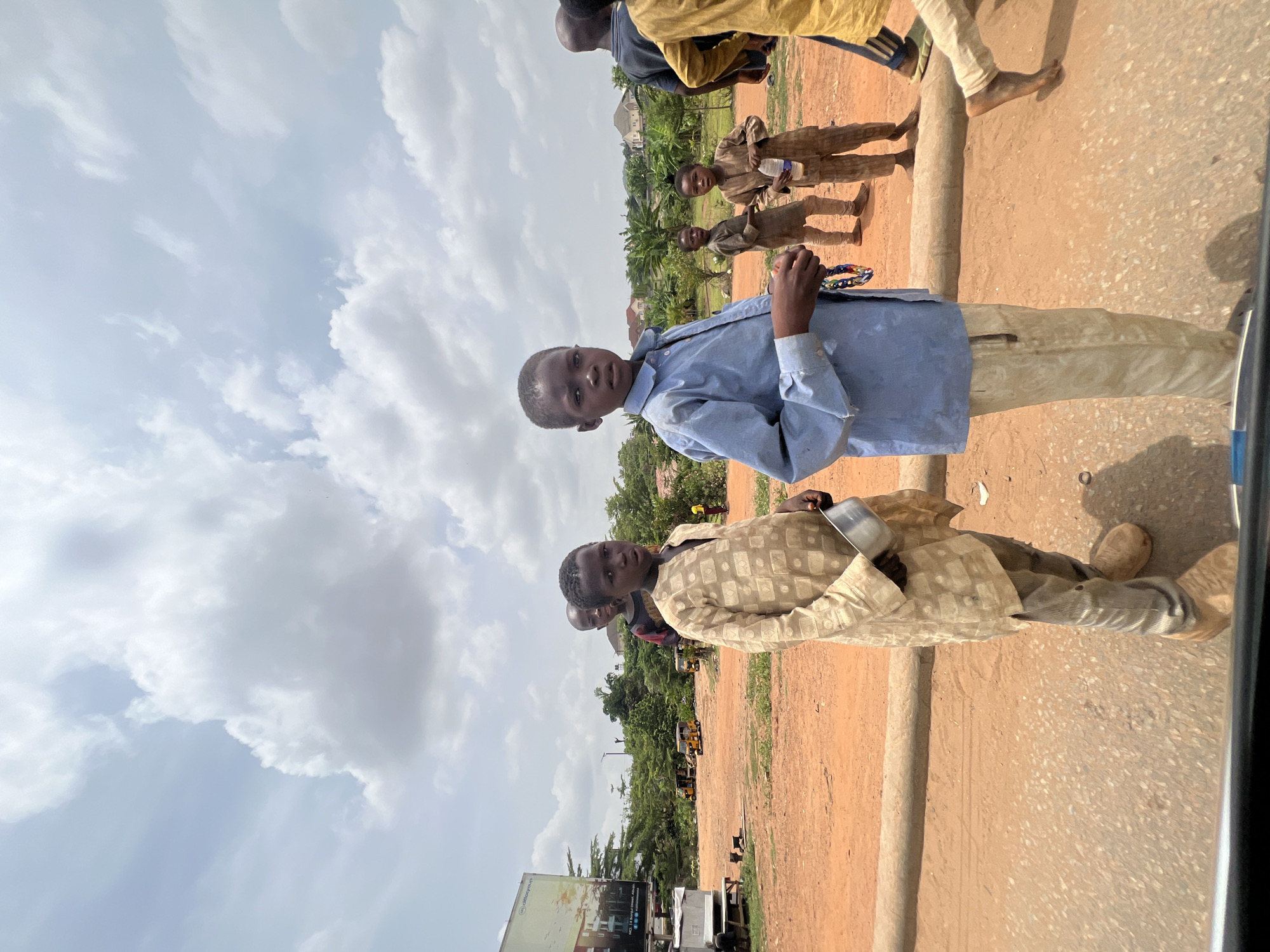 The image captures a street scene in Abuja, Nigeria, under a cloudy sky. A group of young boys stand near a dirt road and a concrete barrier. One boy wears a light blue shirt and tan pants, holding a colorful object. Another boy is dressed in a patterned beige tunic, carrying a metallic container. Other children in simple clothing stand slightly behind them. In the background, there is a billboard, some vegetation, and several yellow auto-rickshaws or "keke napeps". The terrain suggests a less developed or peripheral area within the city.