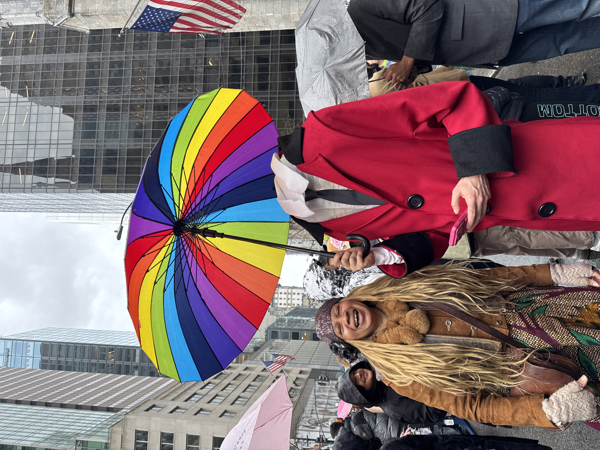 The image captures a street scene in New York City, United States of America, where a woman with long, blonde, dreadlock-style hair is smiling while holding a vibrant rainbow-colored umbrella. She is wearing a brown jacket with a fur collar, a patterned skirt or pants, and textured gloves. To her right, a person in a bright red coat is holding a mobile phone. The umbrella is the focal point, displaying a full spectrum of colors.
In the background, there is a building with a glass facade, reflecting the overcast sky. An American flag is visible near the top of the frame. Several other people are present, some holding umbrellas, suggesting it might be raining or recently has rained. The ground appears wet.