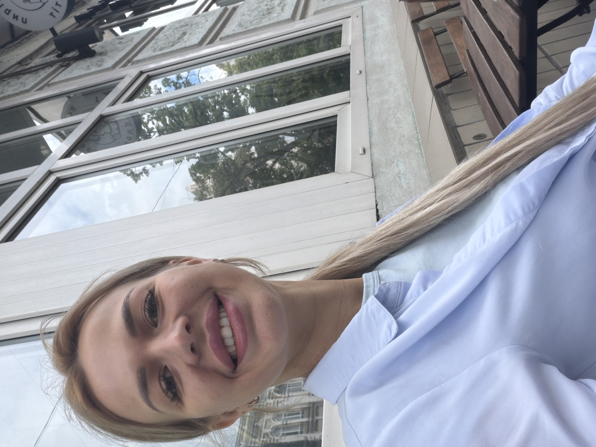 A young woman with blonde hair is taking a selfie in an outdoor setting. She is smiling and has her tongue sticking out. She is wearing a light blue collared shirt. In the background, there are large windows reflecting trees and the sky, suggesting it is daytime and likely pleasant weather. There are also some wooden benches visible. A circular sign with some text is partially visible at the top left of the image. The text on the sign appears to be Cyrillic and includes the word "ТИП". This image was taken in Kyiv, Ukraine.
