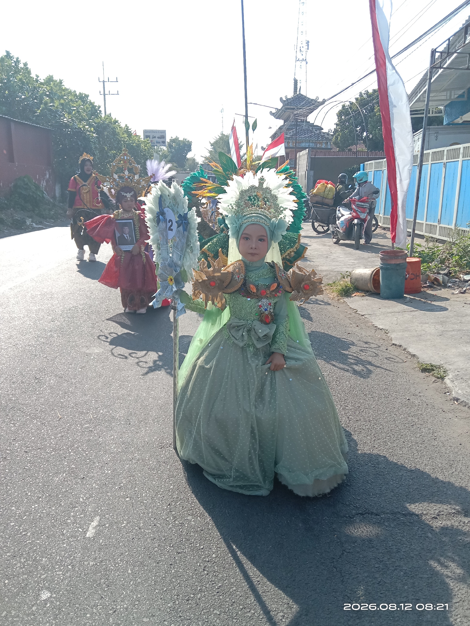 In the city of Gudo, Indonesia, a young girl is participating in a parade. She is dressed in a light green, elaborate traditional costume adorned with sequins and a crown. The dress has a full skirt with a polka dot pattern and is embellished with gold accents on the bodice and shoulders. She is holding a framed photograph of a man. Behind her, other individuals in costumes are also in the procession. One person to her left is wearing a red and gold outfit and carrying a large, decorative staff with white plumes and a number "2" visible on it. Further back, other parade participants and spectators can be seen, along with motorcycles and flags. The parade is taking place on a paved road, with buildings and trees lining the sides. The sun is shining, casting long shadows on the road. The overall atmosphere appears to be festive and celebratory.