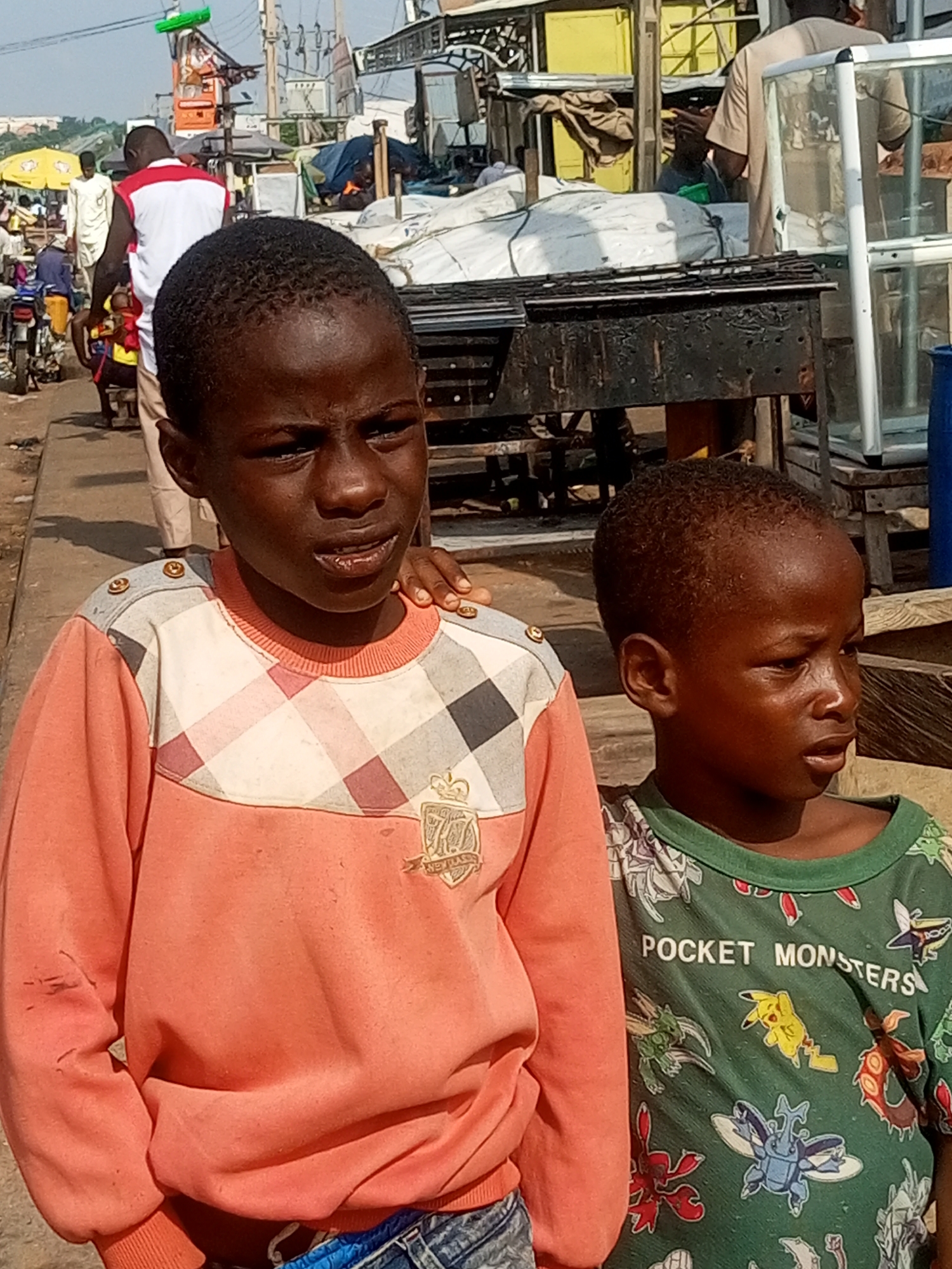 Two young Black males are standing on a paved surface in what appears to be a market area in Tempure, Nigeria. The male on the left is wearing a coral-colored long-sleeved shirt with a checkered pattern on the shoulders and a crest on the chest. He is looking slightly to his left. The male on the right is wearing a green t-shirt with "POCKET MONSTERS" printed across the chest and several cartoon characters. He is looking to his right. In the background, there are various market stalls, including what appears to be a grill or barbecue setup, a glass display case, and a blue barrel. Other people and vehicles are visible in the distance, suggesting a bustling urban environment. The lighting indicates it is daytime.