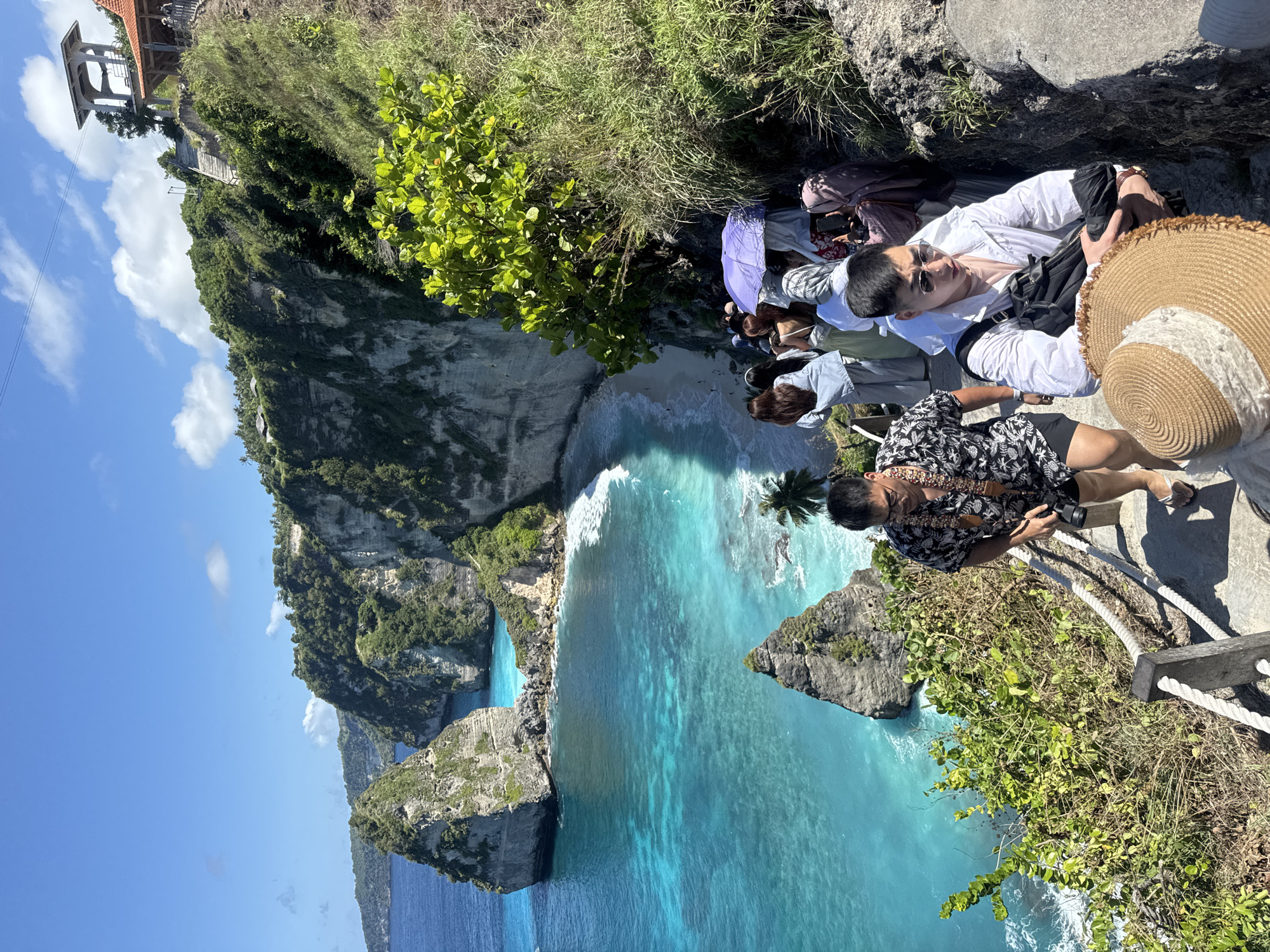 Here is a description of the image:
The image shows a scenic overlook on a cliff in Nusa Penida, Indonesia. The turquoise blue ocean stretches out with rocky formations jutting out of the water. A small beach is visible in a cove between the cliffs. On the edge of the cliff, several people are gathered, taking in the view. One man is wearing a floral shirt and taking photos. Another person is lying down on the rocks with a straw hat beside them. Other people are standing and sitting nearby, with a purple umbrella partially visible. Some greenery and vegetation grow along the cliff edge. A building is visible at the top of the cliff edge to the left.