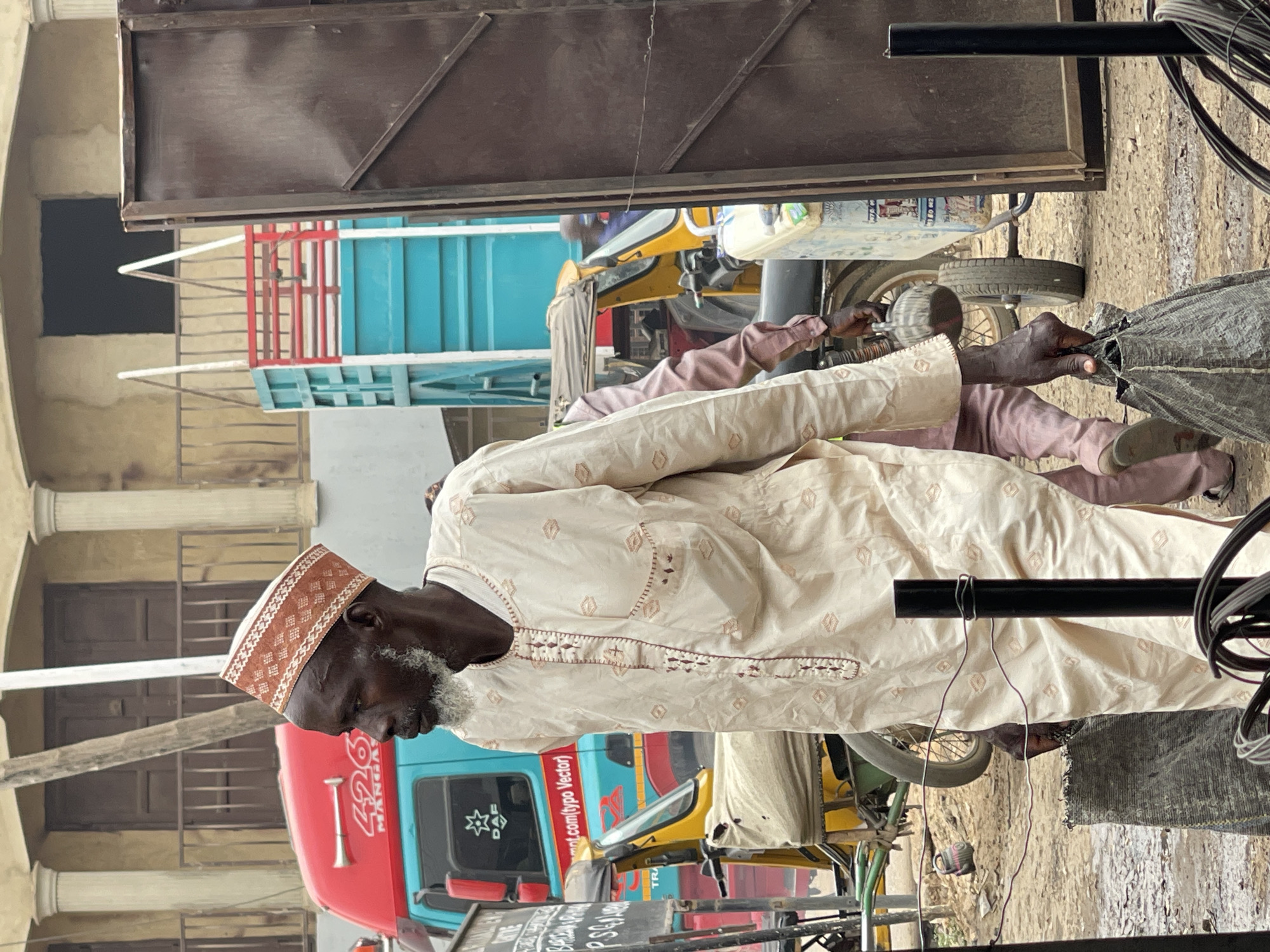 The image depicts a man with gray beard and a brown patterned kufi cap wearing a light brown embroidered traditional garment, interacting with a gray woven-looking bag in the foreground, presumably within the city of Zaria, Nigeria, as suggested by the backdrop. Behind the man, there's a yellow and blue tricycle taxi or keke napep, and a truck with a red cab and blue cargo bed. The building behind the truck features white pillars and dark window openings. Overhead, metal bars and wires extend into the scene. The ground appears to be a dirt or unpaved surface.