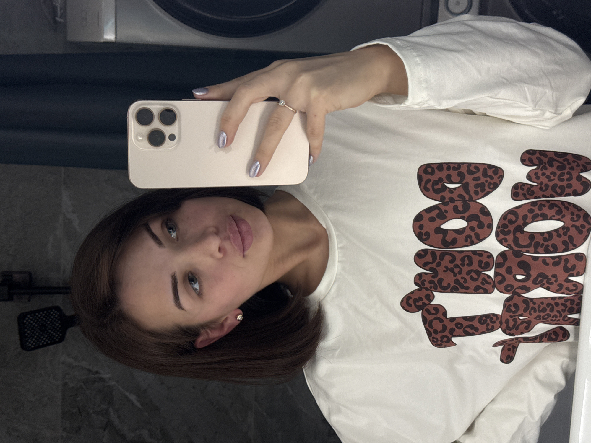 The image shows a woman taking a selfie with a smartphone. She's wearing a white, long-sleeved shirt with a leopard print design. She has medium-length brown hair and is wearing a ring on her left ring finger. The background shows a bathroom setting with a showerhead and a washing machine. The location is Лобаново, Russia.