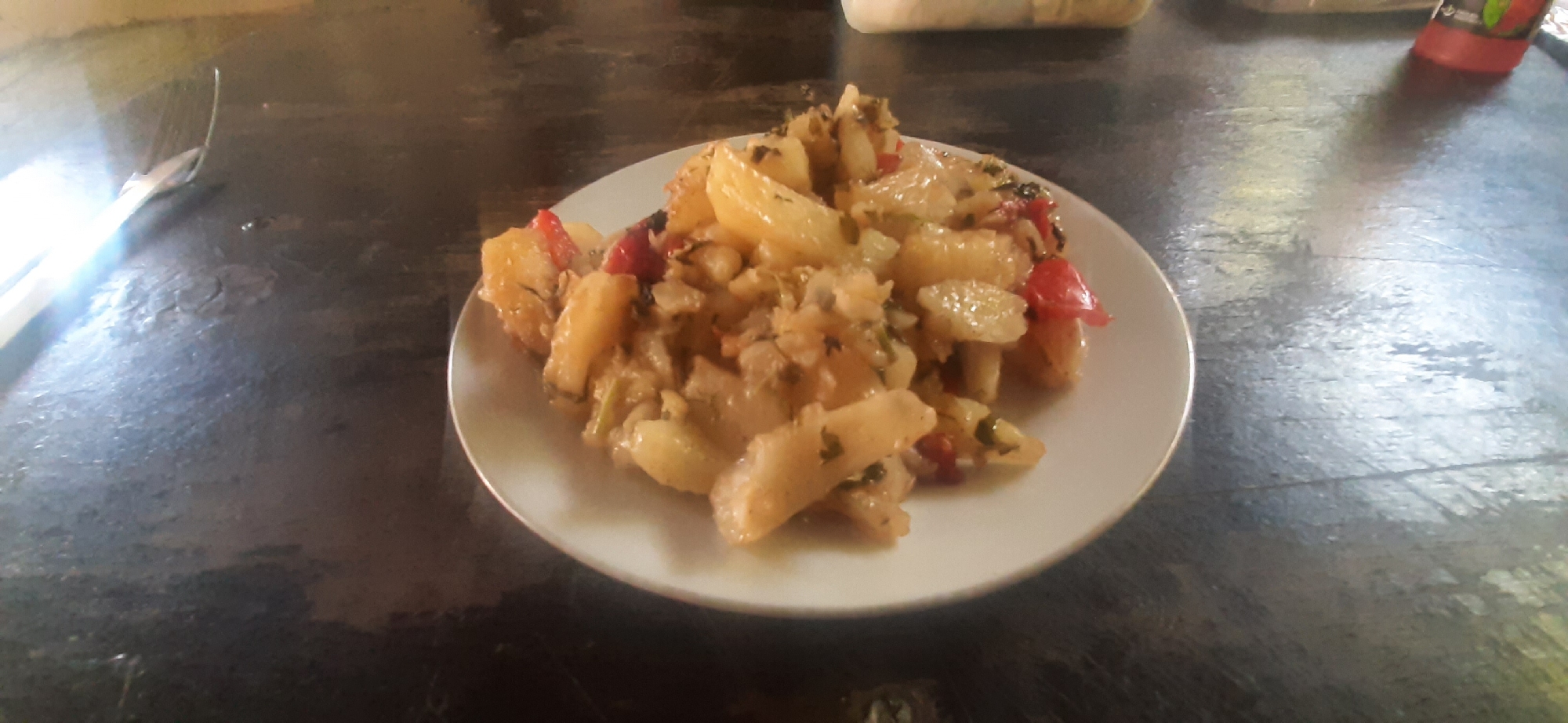 The image shows a close-up of a white plate filled with a potato and red pepper dish, possibly a stew or a sauté. The dish appears to be seasoned with herbs. The plate is situated on a dark, wooden table. To the left of the plate, a silver utensil, possibly a fork or a spoon, is partially visible, reflecting light. In the background, a red bottle with a white cap, likely a condiment or drink, and a blurred white object are present. The scene suggests a casual meal setting. There is no information available about the location in Yessentukskaya, Russia, or any specific people or actions taking place.
