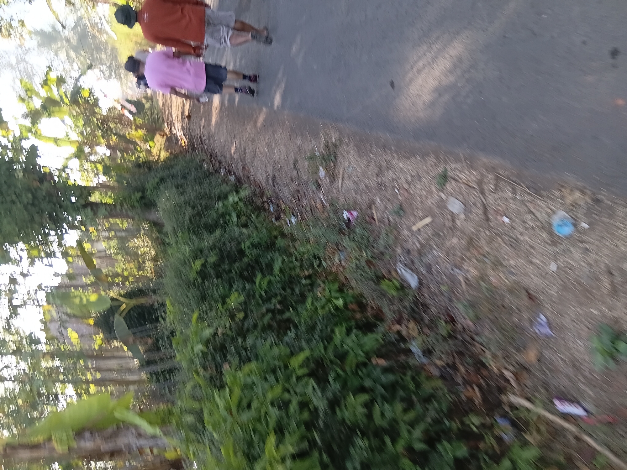 In Tanggul Wetan, Indonesia, two individuals are walking away from the camera on a paved road bordered by lush green foliage. The person on the left wears a pink t-shirt and dark shorts, holding a clear bottle. The person on the right is dressed in an orange long-sleeved shirt and light-colored shorts, also wearing a dark bucket hat. The road is partially shaded by the surrounding trees. Litter, including plastic bags and wrappers, is scattered along the edge of the road and the grassy embankment. The overall scene depicts a casual walk along a rural road in Indonesia.
