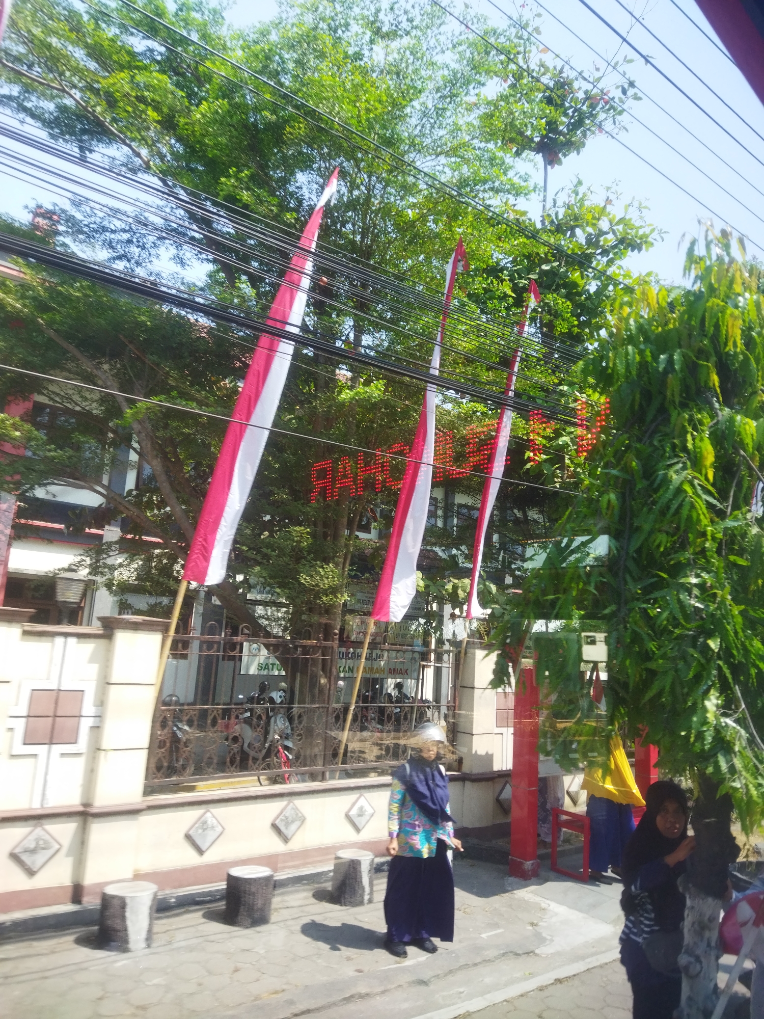 The image shows a street scene in Jombor, Indonesia. In the foreground, there are several concrete cylindrical bollards. A woman wearing a blue hijab and a colorful patterned jacket is standing on the sidewalk, facing left. To her right, another woman in a black hijab is partially visible, leaning against a tree. In the background, there are Indonesian flags with red and white stripes hanging from poles. A building with a white and brown facade is visible behind the flags, and trees with lush green foliage are present throughout the scene. The sky is blue and clear. There are several power lines visible overhead. A red LED sign is partially obscured by trees and flags. A metal fence with decorative patterns separates the sidewalk from the building. Inside the fenced area, motorcycles are parked. The overall impression is of a sunny day in an urban or suburban setting in Indonesia.