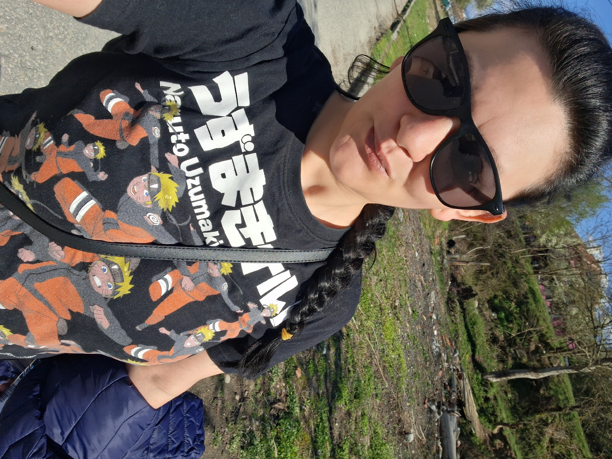 A woman wearing sunglasses and a black t-shirt with images of Naruto Uzumaki and Japanese text is standing outdoors. She has dark hair styled in a braid. She is wearing a navy blue jacket. The location appears to be a semi-rural area near Goce Delchev, Bulgaria, as indicated by the presence of dry grass, dirt, and sparse vegetation in the background. Debris is visible scattered on the ground.