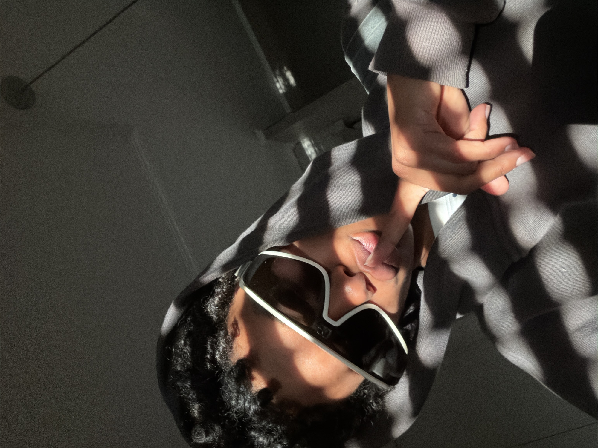 A person wearing a gray hooded sweatshirt and white-rimmed sunglasses is positioned in a room with light and shadow patterns. The person's right hand is raised, with a finger near their lips. The background shows parts of the room including a ceiling light fixture and shelf. The image is tilted. There is no specific information to conclude the location is in Champs-sur-Marne, France.