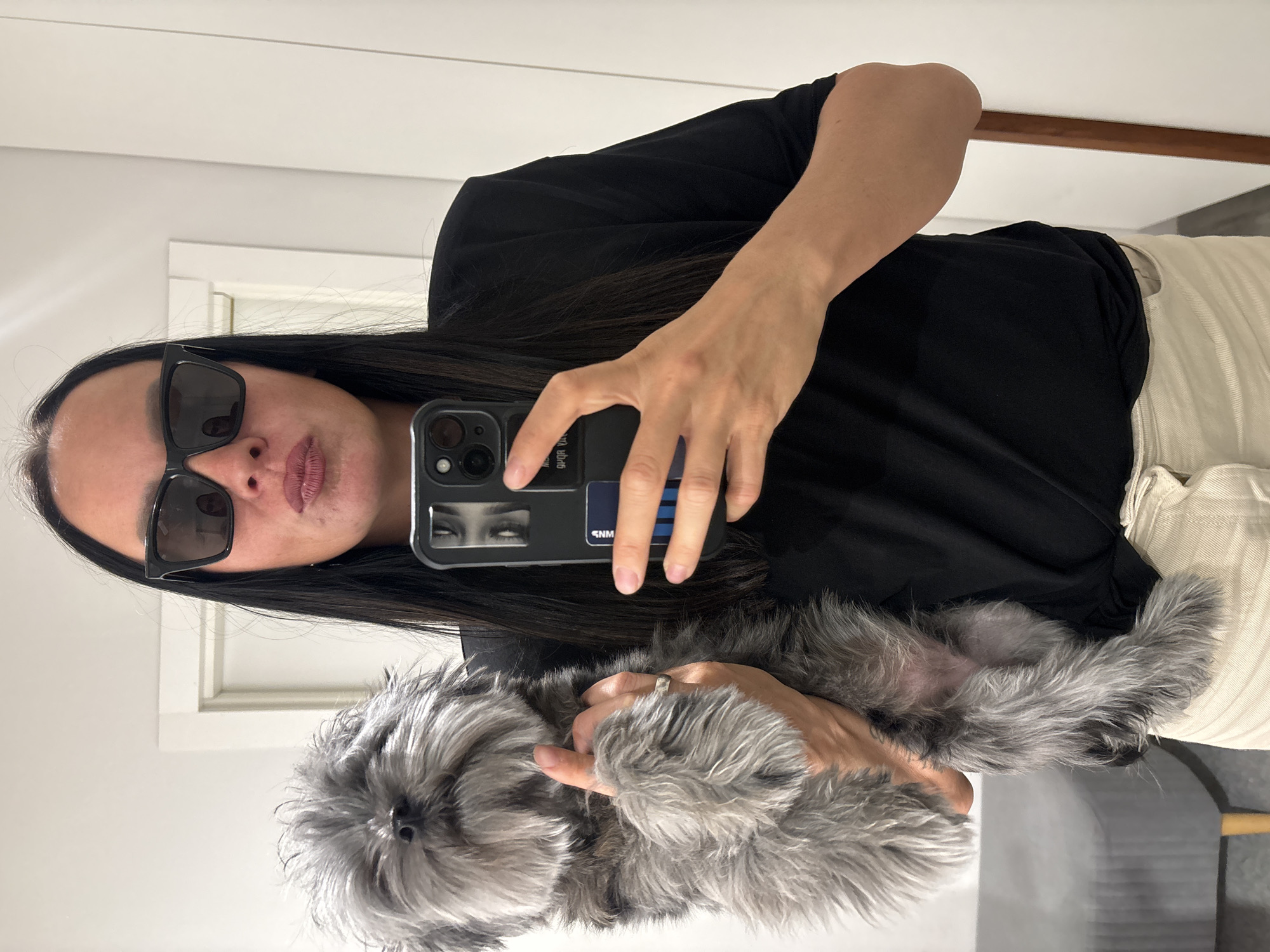 A person with long, dark hair and wearing sunglasses is holding a small, gray, fluffy dog while taking a selfie in front of a mirror. The person is wearing a black top and light-colored pants. A smartphone with a black case is visible. The background includes a white wall with a door frame and a wooden ledge above it. The location is not discernible as being specifically in the city of Chaikovsky, Russia.