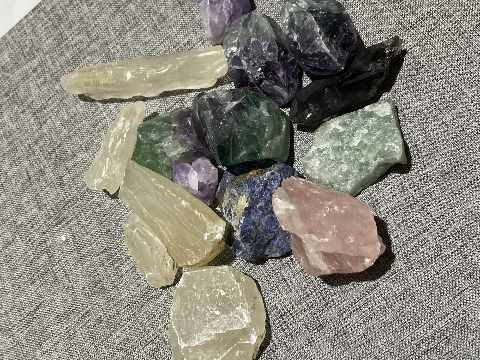 The image displays an overhead view of a scattered collection of various raw and tumbled mineral specimens resting on a light grey, woven fabric surface. The collection includes a range of colors and sizes. Prominent among them are several pieces of clear to pale yellow quartz or calcite, some elongated and others more blocky. There are also multiple chunks of green fluorite, some exhibiting darker green hues, and several deep purple amethyst or fluorite pieces. A single piece of rose quartz is visible, presenting a delicate pink color. A fragment of deep blue sodalite or lapis lazuli with some lighter inclusions is also present. Additionally, there is a distinct black crystal, possibly smoky quartz or tourmaline. The crystals appear unpolished and in their natural, rough state, casting shadows on the textured fabric below. The scene is set in Davao City, Philippines.