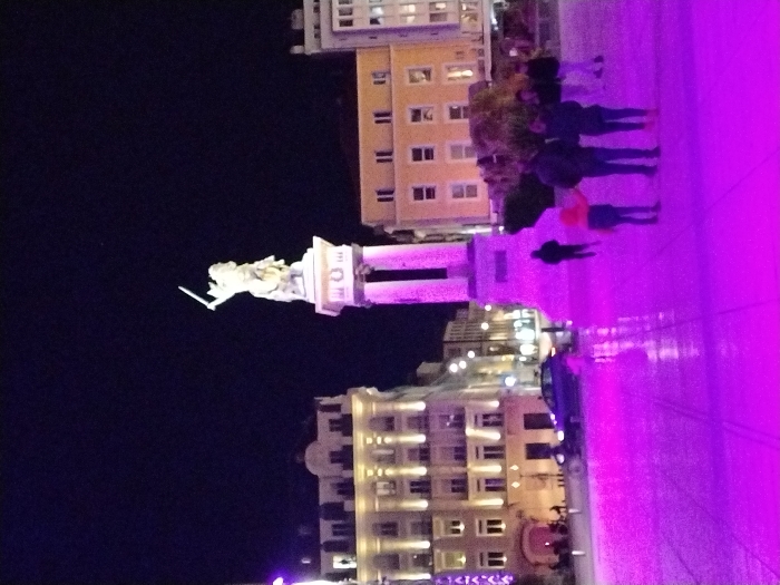 The image displays a nighttime urban scene in Place de Jaude, Clermont-Ferrand, France. The central feature is the equestrian statue of Vercingetorix, depicting him with a sword raised, positioned on a tall, multi-part stone pedestal. The lower portion of the pedestal is illuminated by intense purple light, which also bathes the paved square in the foreground and midground, reflecting off a visibly wet surface.
To the left of the statue, a multi-story building with classical architecture and multiple lit windows is illuminated by warm white external lighting. To the right, a plainer multi-story building also exhibits illuminated windows. A dark-colored sedan is visible in the midground, positioned left of the statue's base.
Approximately seven individuals are present. In the right midground, a group of three individuals—two adults and one child in a bright orange top—stand on the purple-lit pavement. A single, silhouetted individual walks away from the viewer in the left midground. Additional indistinct figures are visible further in the background. The sky is dark and clear, indicating nighttime conditions.
