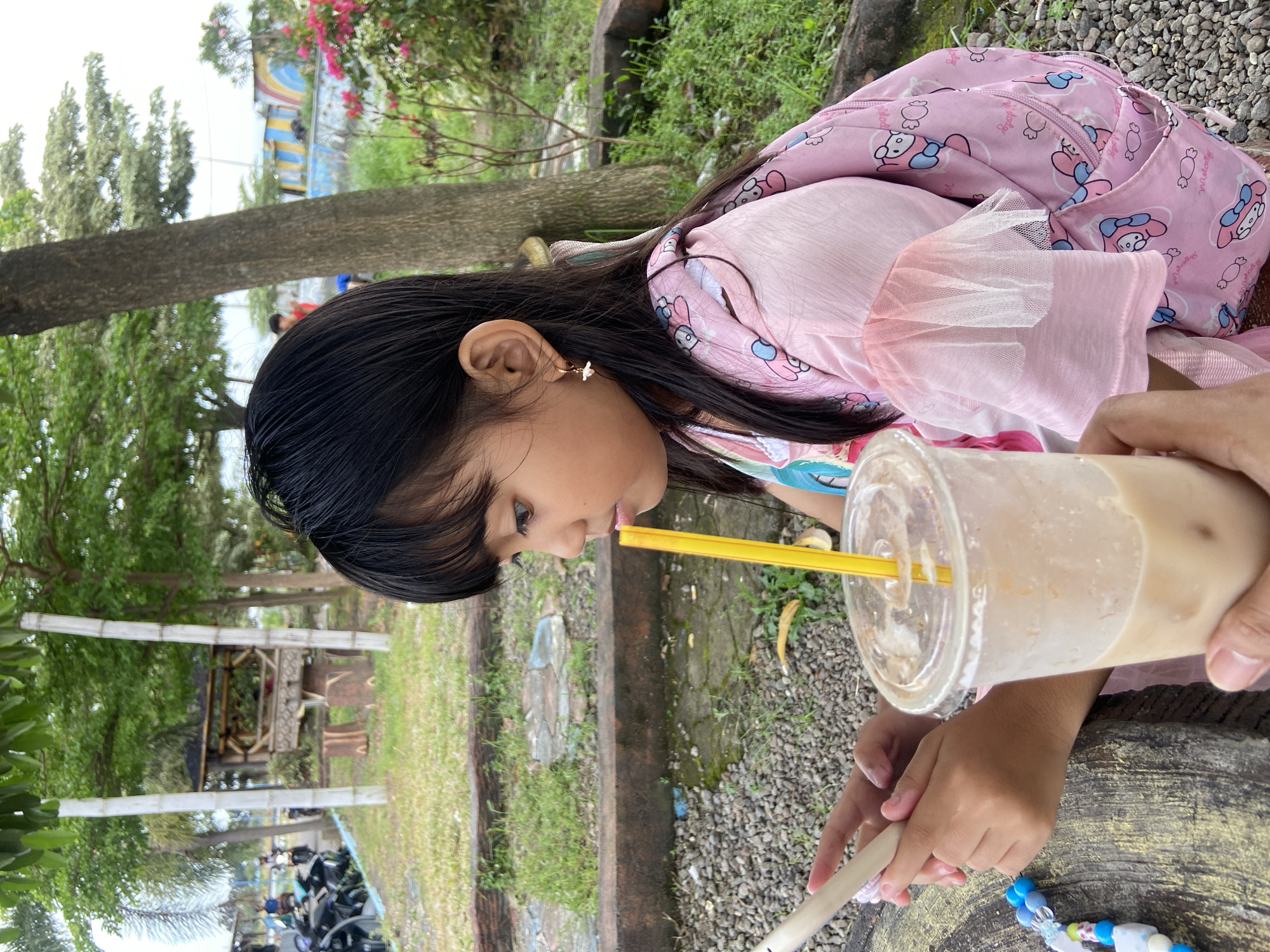 A young girl is drinking from a plastic cup with a straw, holding a white object, sitting outside in Kalitengah, Indonesia. She has dark hair and is wearing a pink shirt and backpack. The setting appears to be a park with trees, green grass, and a gravel surface. Motorbikes are visible in the distance.