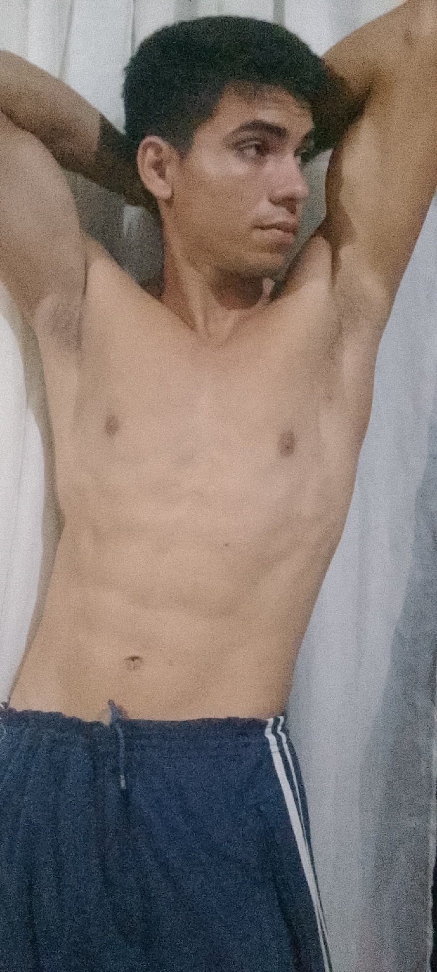 A shirtless, light-skinned young man with dark hair is posing indoors. He is wearing dark blue athletic shorts with white stripes down the sides. His hands are behind his head, arms raised, and he is looking off to the side. The background appears to be a white curtain or wall. The exact location in Juan Griego, Venezuela is not discernible from this image.