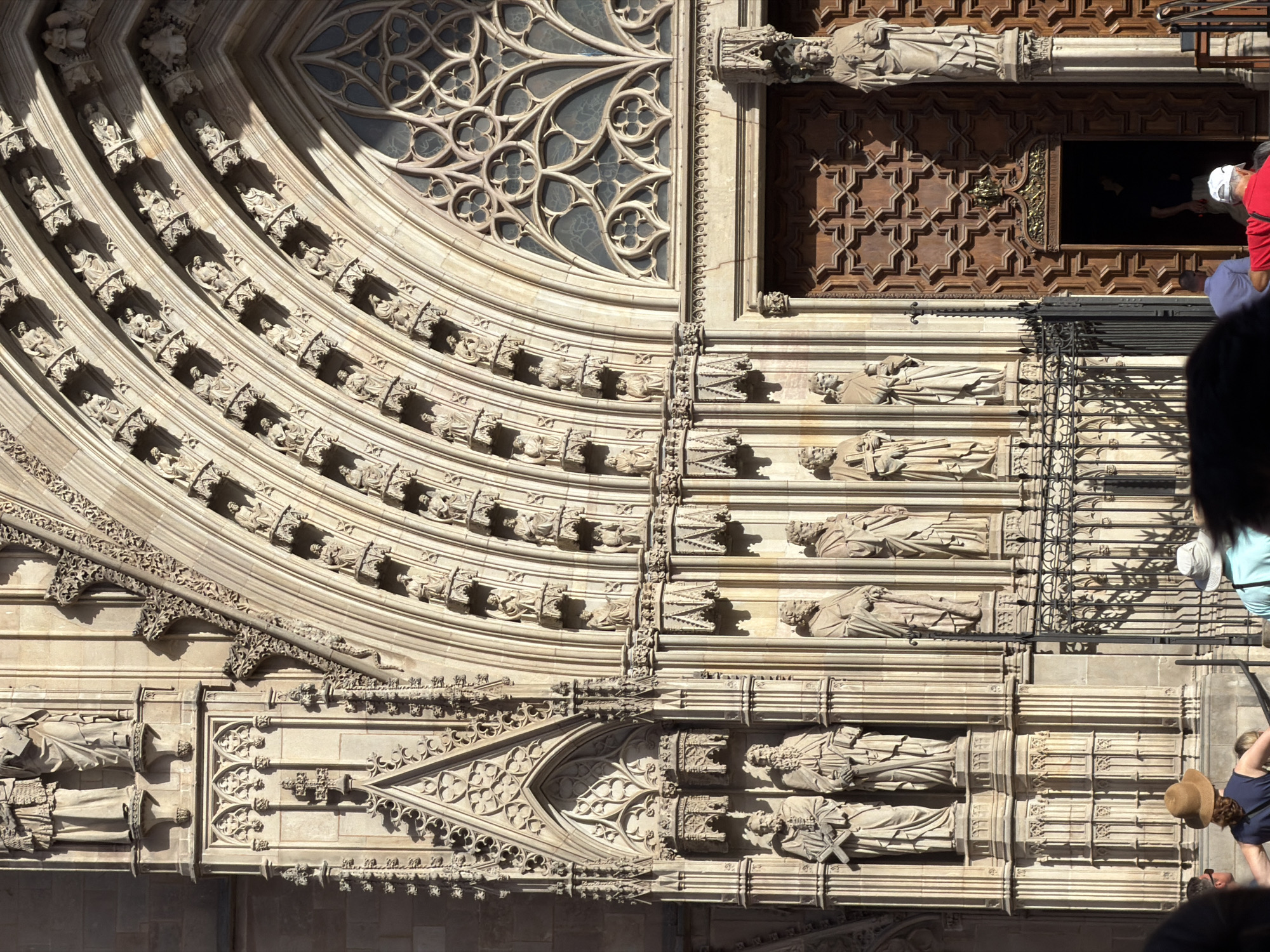 This media file shows a close-up view of a section of the Barcelona Cathedral in Spain, specifically focusing on its ornate facade. The architectural style is Gothic, characterized by intricate carvings, pointed arches, and decorative tracery.
The main subjects are the architectural details of the cathedral, including rows of sculptures of figures in niches, which appear to be saints or religious figures. These sculptures are carved from stone and are weathered, suggesting the building's age. The facade also features a large, elaborate rose window with geometric patterns, though only a portion of it is visible. Beneath the window is a grand wooden door with a complex star-shaped pattern, set within a stone portal.
Several people are present in the scene, mostly at the bottom and right edges, suggesting they are tourists observing the cathedral. One person in the upper right is wearing a red shirt and a white hat, and another person below them wears a blue shirt. Further down, a person wearing a hat is bent over, looking at something. Their presence indicates that this is likely a daytime scene, given the sunlight and shadows cast on the facade. The weather appears sunny, contributing to the distinct shadows that highlight the depth and texture of the carvings.
Notable details include the fine craftsmanship of the stone carvings, the geometric patterns of the rose window and the wooden door, and the presence of iron railings that likely delineate an entrance or a viewing area. The scale of the architectural elements and the detailed ornamentation convey a sense of grandeur and historical significance. There is no visible text in the media file.