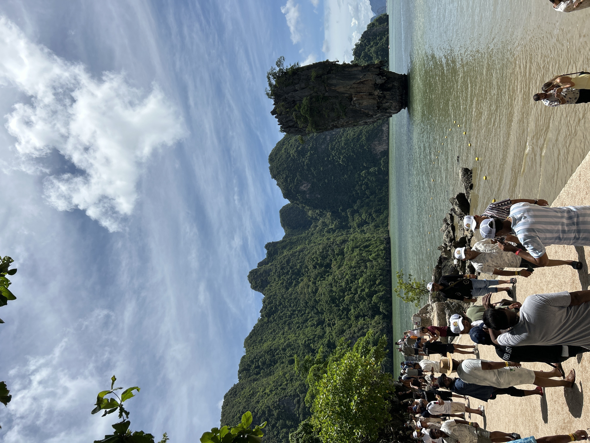 This image depicts the iconic James Bond Island in Krasom, Thailand. A large group of tourists are gathered on a sandy and paved pathway along the coast, observing the distinctive limestone karst rock formation that rises dramatically from the turquoise water. The island is covered in lush green vegetation, with a small cluster of trees growing on its peak. The sky above is partly cloudy, with a mix of blue sky and white clouds. The water is calm and reflects the surrounding greenery and sky. Some people are standing, while others are sitting or walking. The atmosphere appears to be one of leisure and sightseeing.