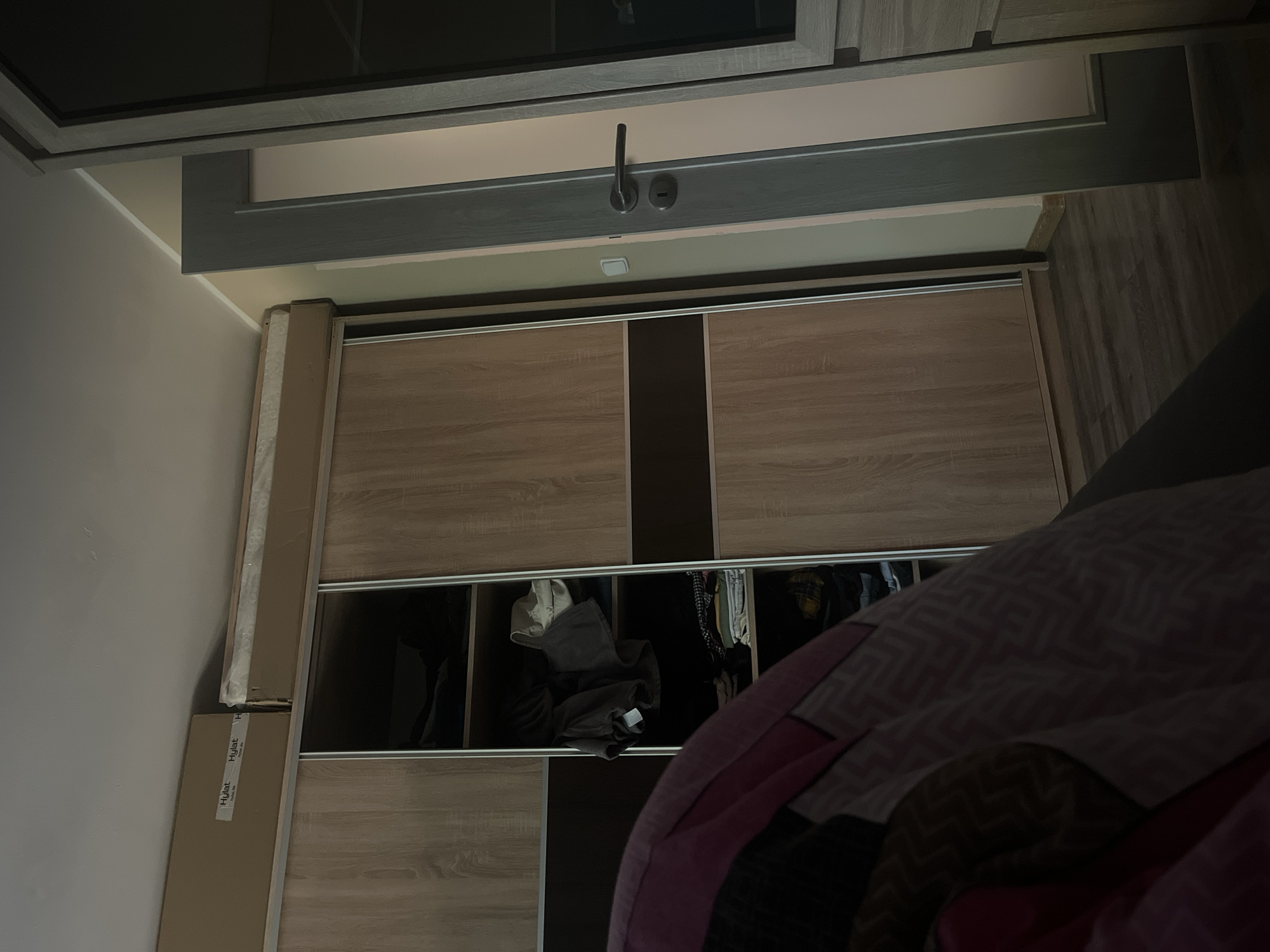 The image shows an interior view, potentially within a residential building in Břeclav, Czechia, given the location specification. The main subject is a closet with sliding doors, which appears to be partly open, revealing its contents. Clothes are visible inside the closet. The closet is made of a wood-like material with dark vertical accents. To the left of the closet stands a cardboard box. The top of the image shows another part of the building's architecture, likely another cabinet or shelf structure. A large patterned blanket partially obscures the view in the foreground on the right. No people are visible in the image.