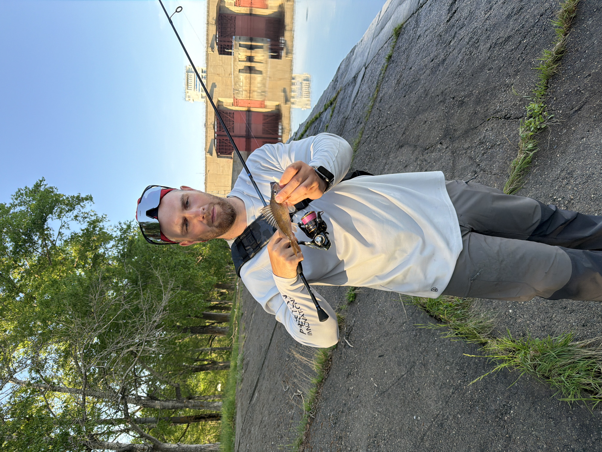 The image shows a man holding a small fish on a fishing rod. He is standing on a concrete embankment with grass growing in the cracks. In the background, a building or structure is reflected in the water. The location appears to be the Chaikovsky dam in the city of Chaikovsky, Perm Krai, Russia. The man is wearing sunglasses, a long-sleeved shirt, and a watch. He has a fishing reel attached to his rod. The environment suggests a sunny day.