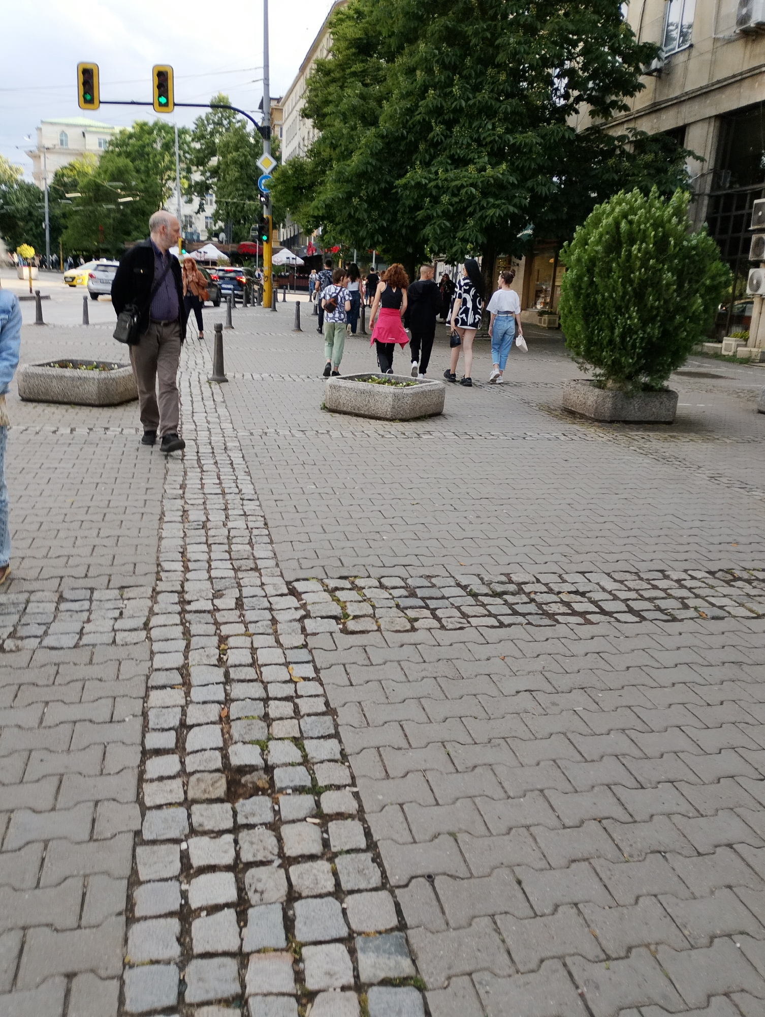 The image captures a vibrant urban street scene from a low-angle perspective, focusing on a paved sidewalk. The foreground features a pedestrian area composed primarily of interlocking grey concrete pavers, with a distinct central strip of older, irregular cobblestones running down the middle.
Several people are visible, walking along the sidewalk. In the immediate foreground, a man with gray hair, dressed in a dark jacket, brown pants, and carrying a black shoulder bag, walks towards the left side of the frame. Further back, a group of individuals walks away from the viewer, including a woman in a black top and red jacket, a person in a black shirt, a woman in a black and white patterned top and shorts, and others in casual wear like blue jeans and white tops.
The background reveals a city street with vehicles, including a yellow taxi and a dark car. Overhead, two distinctive rectangular yellow traffic light casings display green lights. A standard traffic light further down also shows green. Street signs, including a diamond-shaped "priority road" sign and a blue circular "keep left" sign, are visible near an intersection.
The street is flanked by tall, leafy green trees and multi-story buildings. A neatly trimmed green bush sits in a concrete planter on the right side of the sidewalk, with other similar planters holding small plants elsewhere. Bollards line parts of the sidewalk. The sky appears overcast, suggesting daytime with soft, diffuse lighting. No legible text is visible on any signs or buildings. The scene depicts a routine moment in a bustling city environment.
