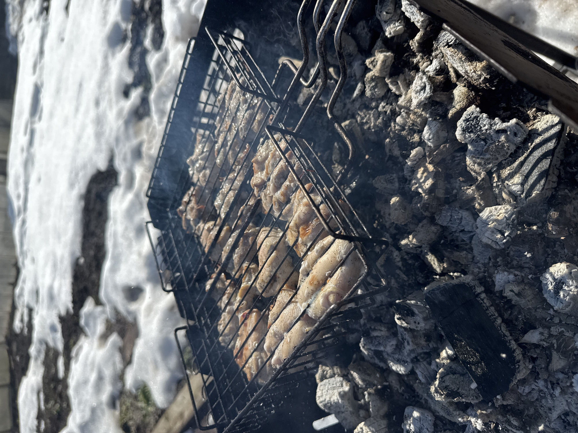 The image shows a grill with meat cooking over coals in the city of Фоки, Russia. Snow is visible in the background. The grill is black metal and contains pieces of meat, possibly chicken, arranged inside. The coals beneath are gray and white ash, with remnants of unburned wood. Smoke rises from the cooking meat. The setting appears to be outdoors, judging by the presence of snow and natural light.