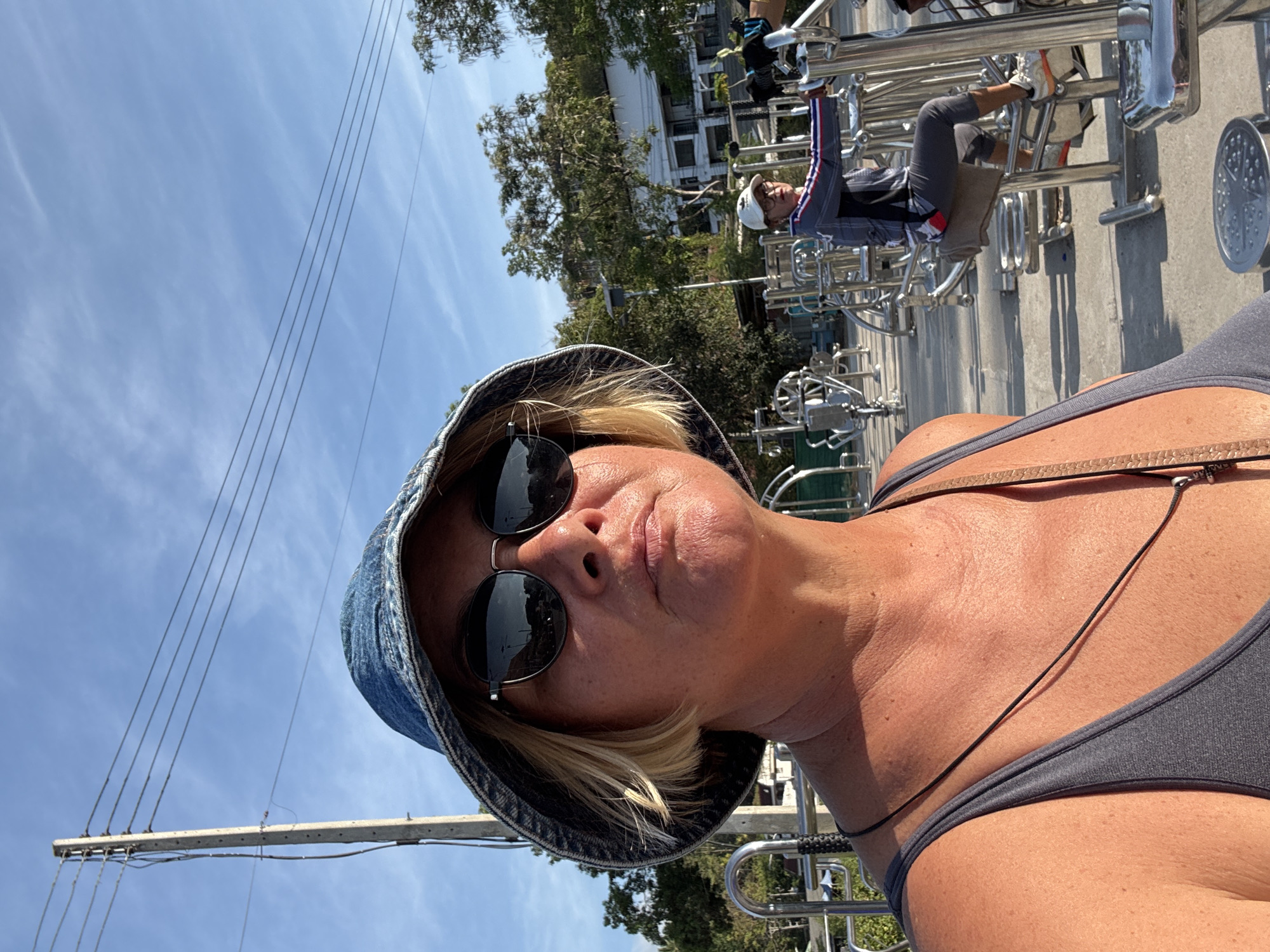 A medium-shot, rotated photograph captures a woman in the foreground with a man partially visible in the background. The woman wears a denim bucket hat and sunglasses, and a gray tank top. A thin necklace hangs around her neck. The background features a man exercising on what appears to be outdoor exercise equipment, constructed of metal bars. Behind the man is a view of trees and a partial view of a light-colored building. Power lines are visible against a blue sky. The location is identified as บ้านบางรักษ์, Thailand.