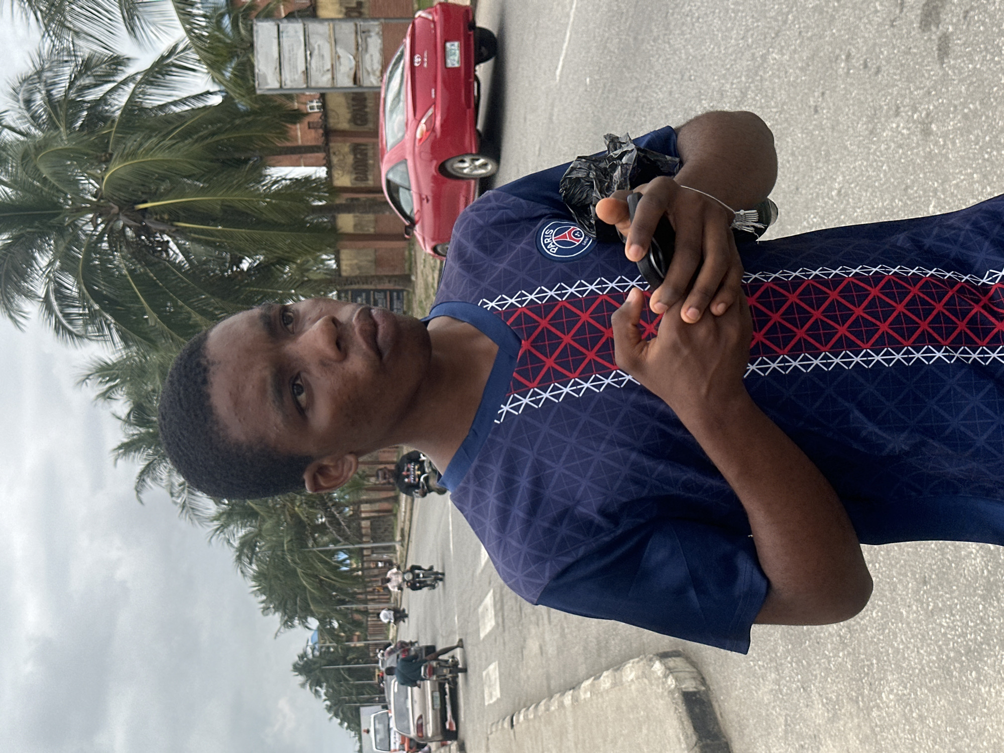 A young Black male stands on an asphalt road in Badagry, Nigeria, looking to his right with a neutral expression. He is wearing a navy blue t-shirt with a red and white geometric pattern on the front. His hands are clasped together in front of him, holding a black smartphone and a crumpled black plastic bag. Behind him, palm trees line the street, and several vehicles, including motorcycles and cars, are visible in motion. The sky is overcast.