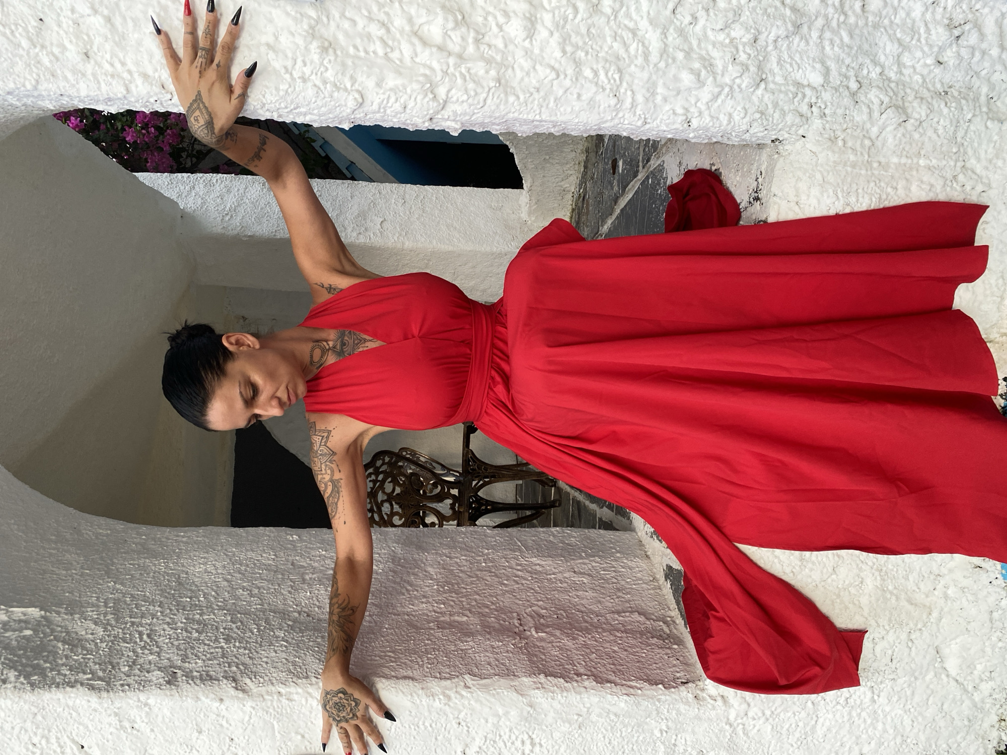 A woman wearing a long red dress leans against a white wall. She has tattoos on her arms and hands, and her nails are long and black with one red nail. The location appears to be Phu Thuy Ward, Vietnam, judging from the architecture visible behind the wall. A chair is visible in the background.