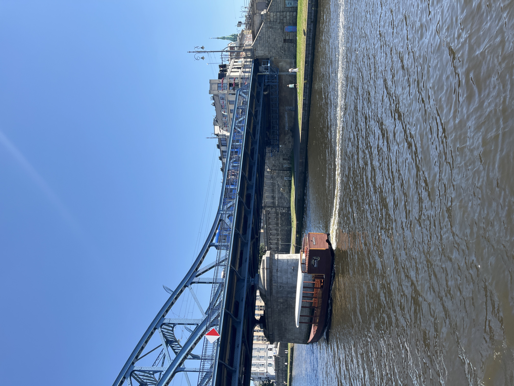 An exterior, eye-level, daylight, medium shot captures a brown tour boat with a white canopy moving right to left on a wide river, creating a foamy wake. The boat has a covered section with visible wooden seating and text on its stern that reads "BAVVA KRAKOW.PL". In the background, a large, blue, steel truss bridge with train tracks running across it spans the river. The bridge supports are substantial stone structures. On the right bank of the river, a grassy embankment leads up to a stone retaining wall and buildings, including a church spire. Two people are visible walking on the grassy embankment. The sky is clear and blue.