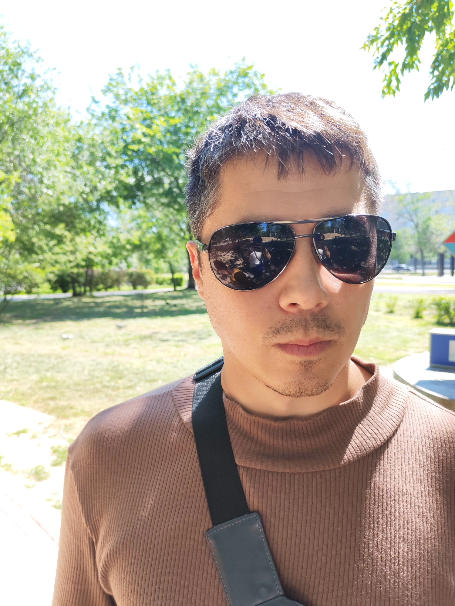 This is a close-up, selfie-style photograph of a person wearing sunglasses and a brown ribbed turtleneck shirt. The subject is looking directly at the camera with a neutral expression. The sunglasses are aviator-style with dark, reflective lenses. A black strap from a bag or camera is visible crossing the subject's shoulder. The background appears to be an outdoor park setting with green grass and trees, suggesting a daytime scene with bright sunlight. The lighting indicates it is likely a sunny day, and the dappled sunlight on the subject's hair suggests the presence of foliage overhead. The overall mood is casual and unposed. No visible text is present in the image.