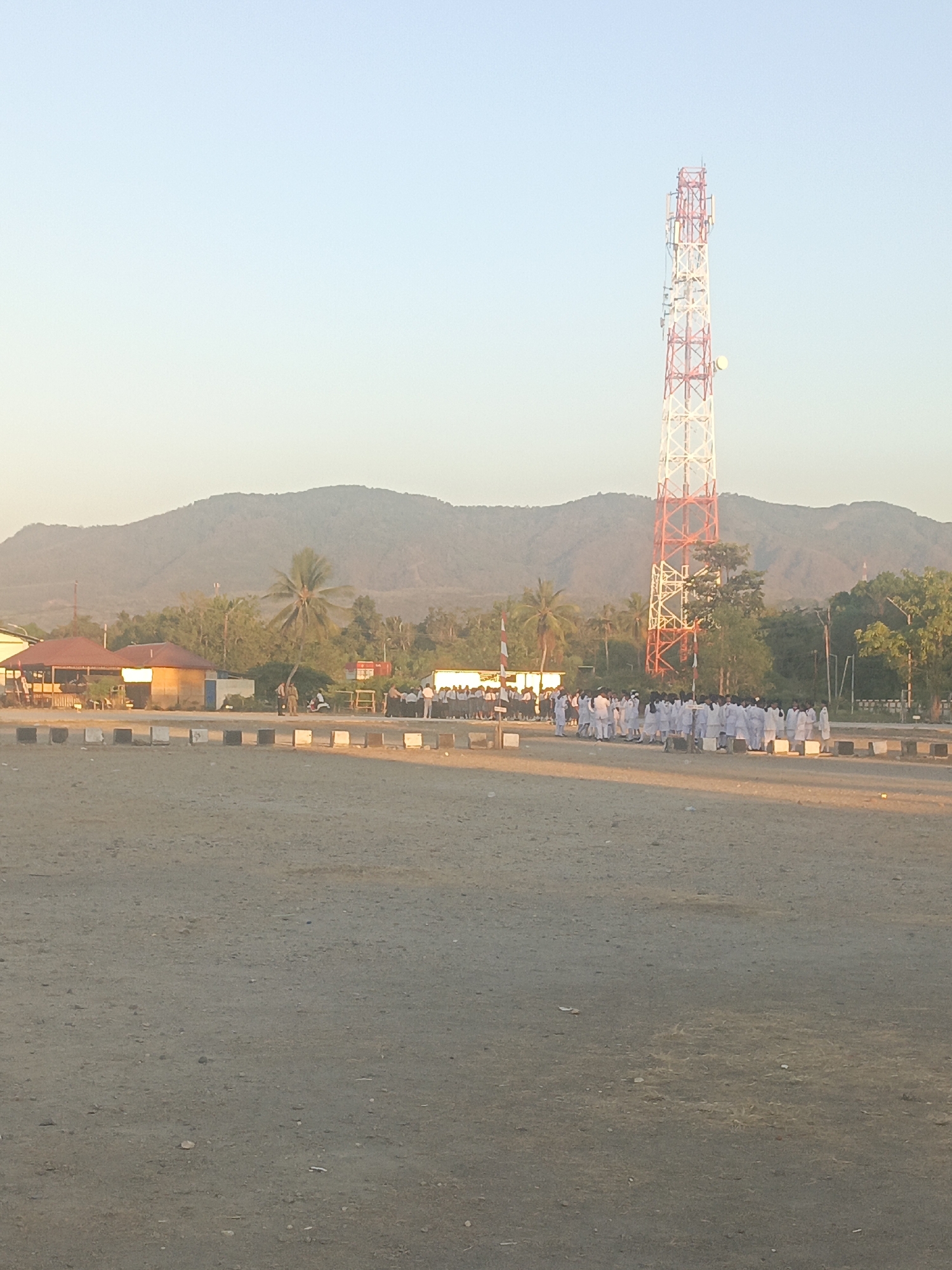 The image captures a wide view of a dusty, open area in Lewoleba, Indonesia. Dominating the background is a tall, red and white telecommunications tower. Behind the tower, a large, gently sloping mountain range is visible under a clear, pale blue sky. In the mid-ground, several low-lying buildings with reddish roofs are situated, some appearing to be commercial establishments or community buildings. Adjacent to these buildings, a group of people, predominantly young individuals dressed in white uniforms, are standing in formation. Some appear to be in the process of assembling or dispersing. A few palm trees and other foliage are scattered throughout the scene, suggesting a tropical or subtropical environment. The foreground is characterized by a flat, barren expanse of dirt and small stones. The overall lighting indicates it is either early morning or late afternoon, with a warm glow illuminating the scene.