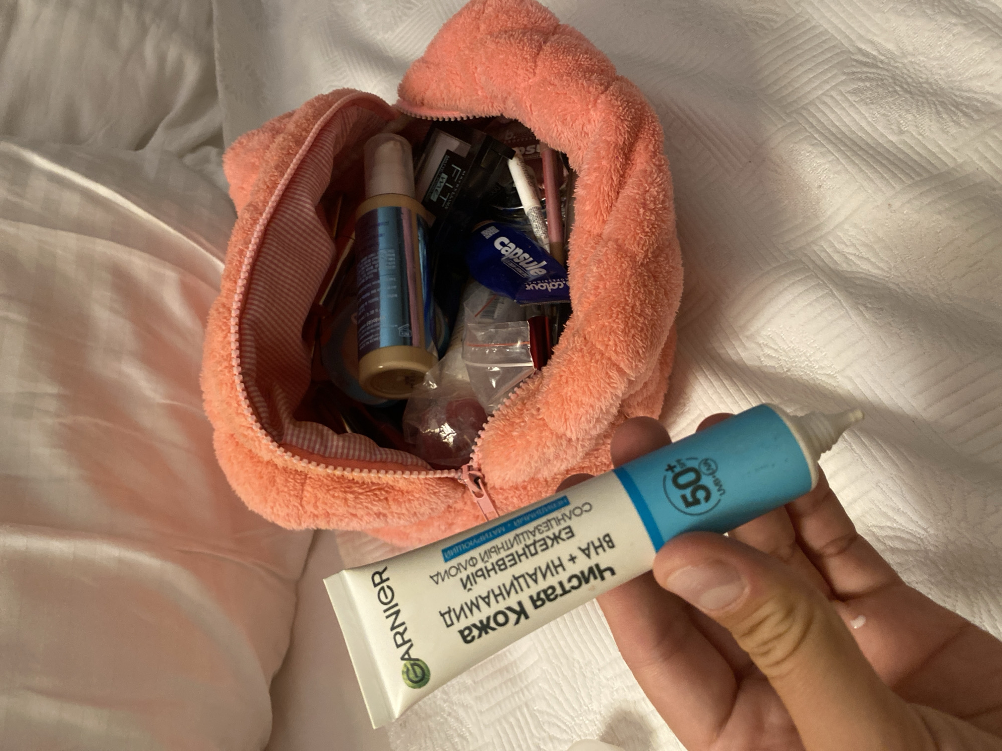 A hand is holding a tube of Garnier "Chistaya Kozha" (Pure Skin) moisturizer with SPF 50. The tube is blue and white with Russian text. The hand has a small amount of white cream on the index finger. Behind the hand, an open, peach-colored, fuzzy cosmetic bag is spilling out various makeup items, including lipstick, pencils, and small bottles. The cosmetic bag and its contents are resting on a white, textured surface, possibly a bedspread. The scene appears to be indoors, with soft lighting. There is no indication of the city of Aksu, Turkey.